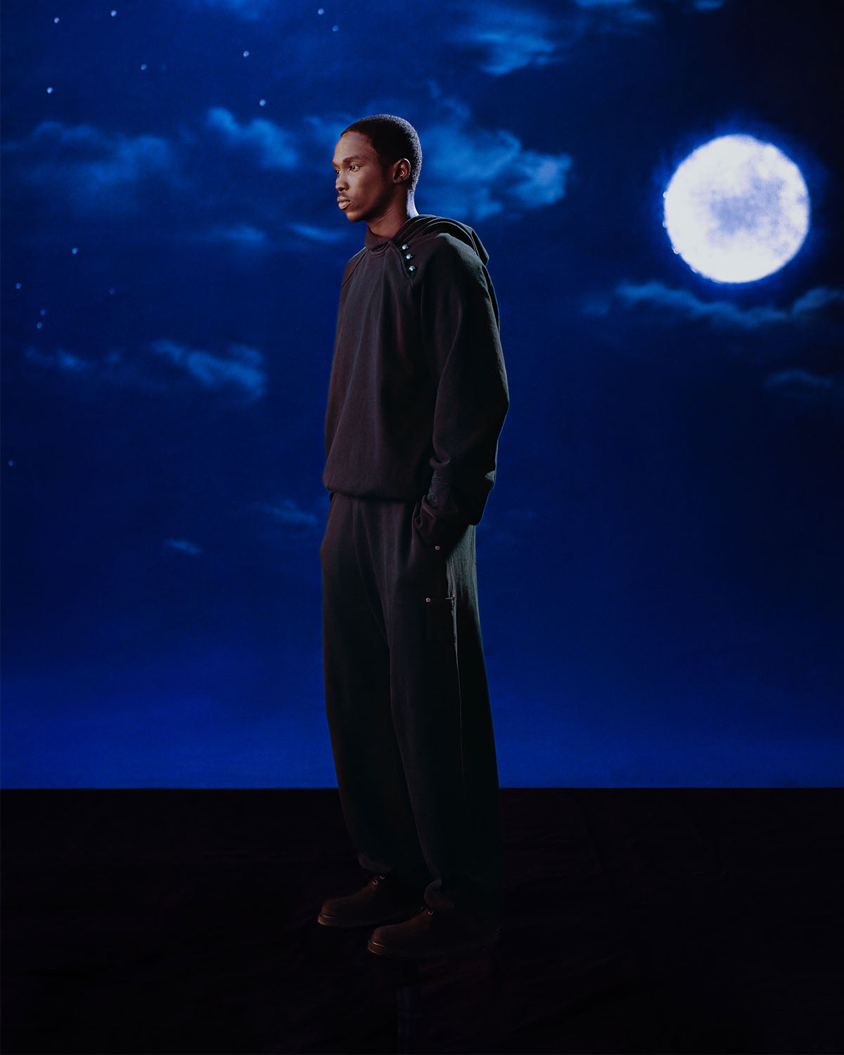 A man stands before a moon, wearing Sundae School’s FADE BLACK HANBOK HOODIE featuring an asymmetric hood closure and signature stash pocket.