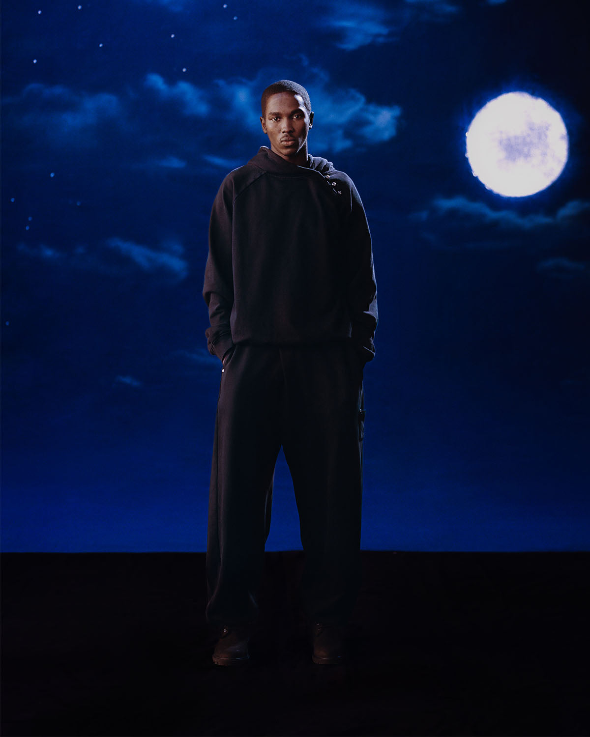 Someone wearing the Sundae School FADE BLACK HANBOK HOODIE, featuring a loose fit and asymmetric hood closure, stands in front of a night sky with scattered clouds and a bright full moon.