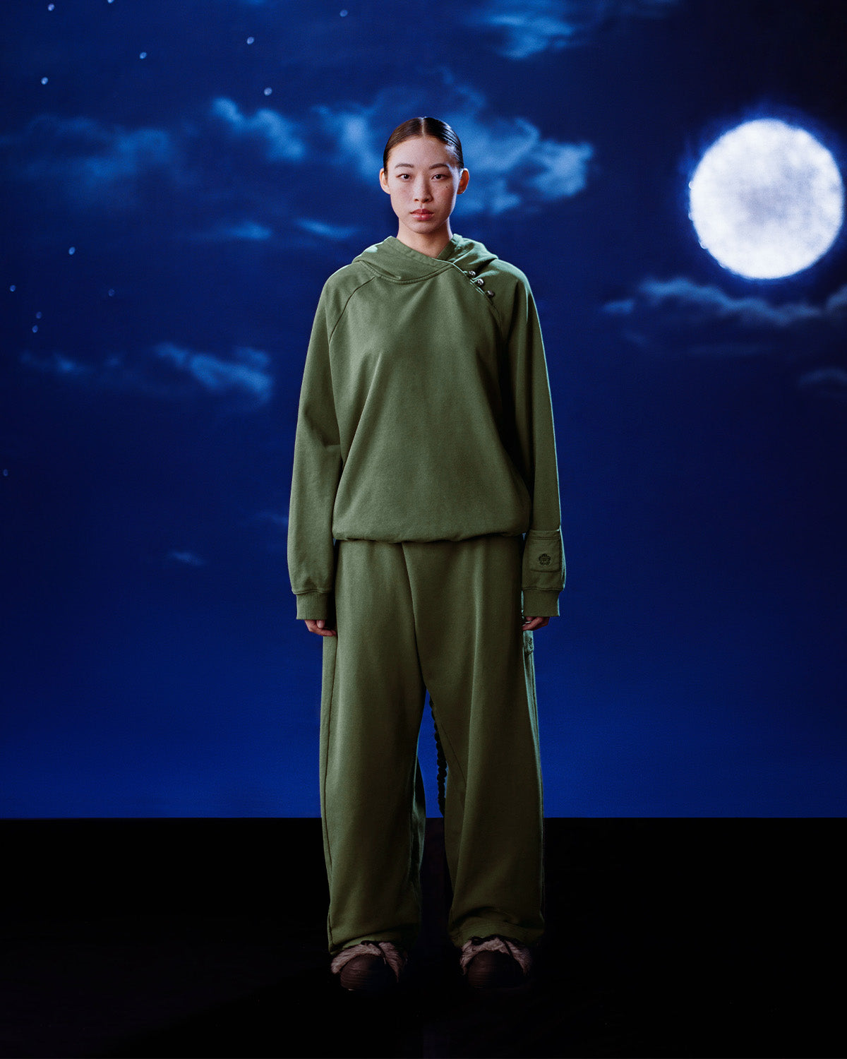 A person stands under a cloudy night sky with a full moon, wearing Sundae School’s oversized KHAKI HANBOK HOODIE and matching pants, hands by their sides and a neutral expression.