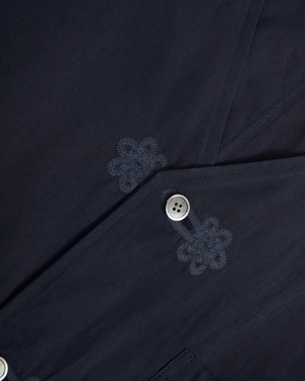 Close-up of the MAEHWA COLLARLESS SHIRT by Sundae School in dark navy, featuring two buttons and intricate Maehwa embroidery near the buttonholes. The smooth, finely detailed fabric enhances the refined aesthetic of this unisex mid-layer.