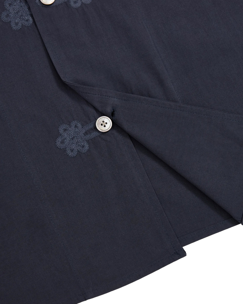 Close-up of the Sundae School MAEHWA COLLARLESS SHIRT, crafted in dark blue linen without a collar and accented by white buttons. Near the buttonholes, delicate Maehwa embroidery adds a unique touch. The slightly open weave reveals an inner layer, making this unisex shirt an ideal mid-layer for versatile styling.