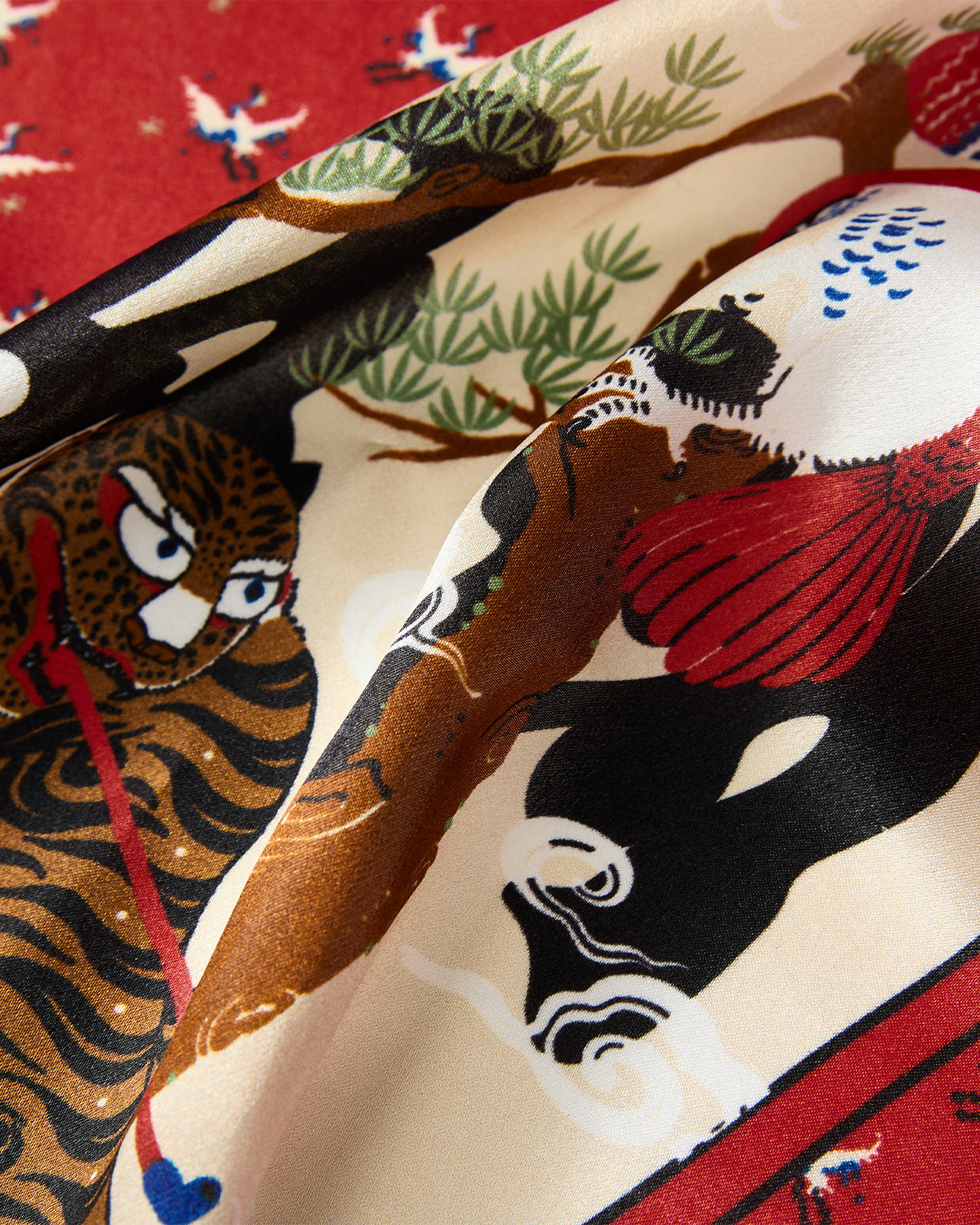 A close-up of the MINHWA BANDANA by Sundae School, featuring colorful Korean folk art-inspired patterns with a tiger, bird, and stylized plants on red and cream silk—an elegant, vibrant accessory perfect as a luxury gift.