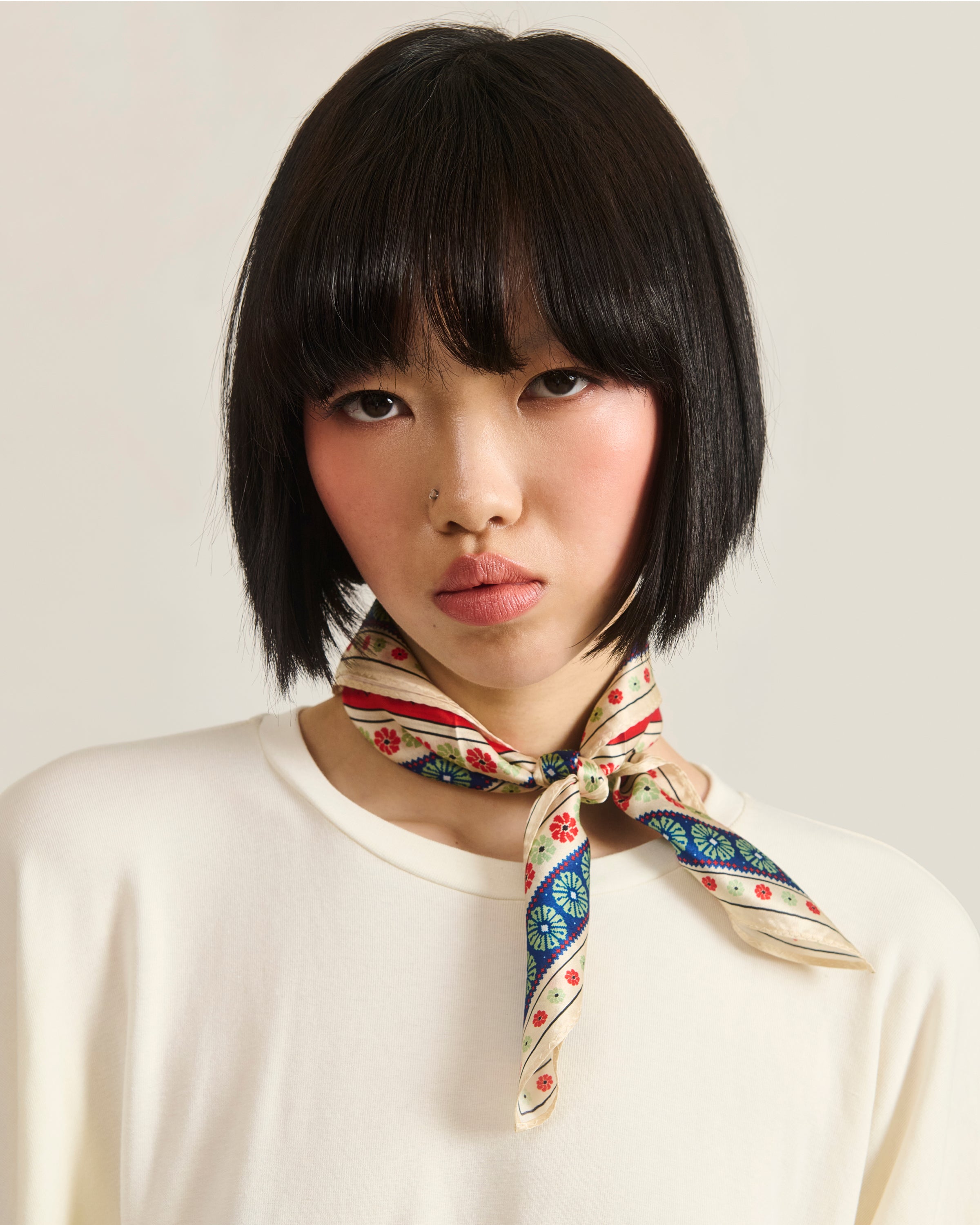 A woman with short black hair and bangs wears a cream top and the MINHWA BANDANA by Sundae School, inspired by Korean folk art, tied around her neck. She looks directly at the camera with a neutral expression against a light background.