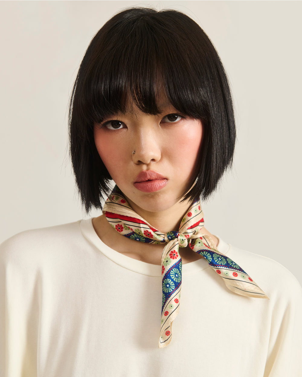 A woman with short black hair and bangs wears a cream top and the MINHWA BANDANA by Sundae School, inspired by Korean folk art, tied around her neck. She looks directly at the camera with a neutral expression against a light background.