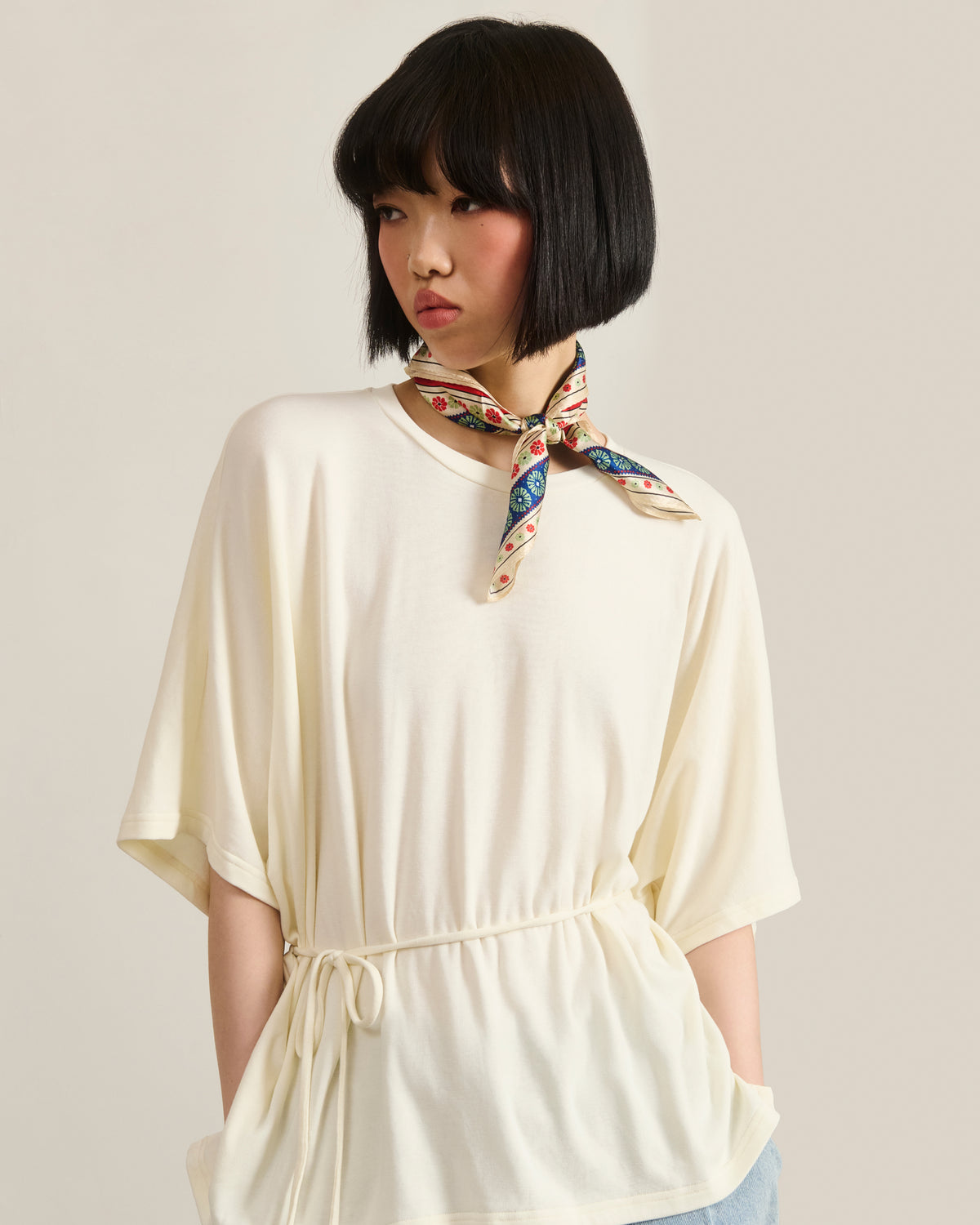 A person with a short black bob wears a loose white top with a waist tie and the Sundae School MINHWA BANDANA, inspired by Korean folk art, standing against a plain light background.