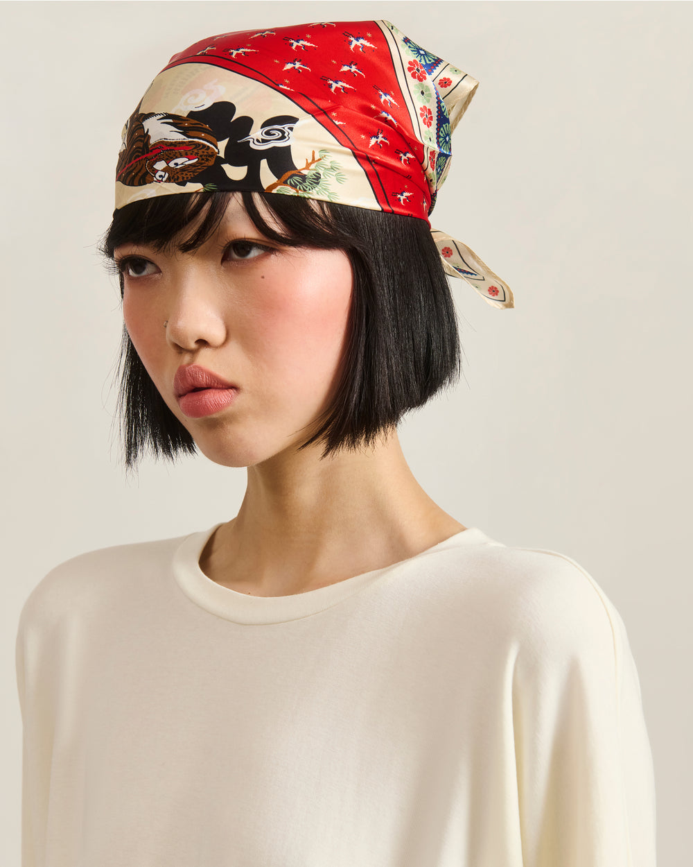 A woman with a short black bob wears Sundae School’s MINHWA BANDANA, a red silk scarf inspired by Korean folk art, tied around her head, paired with a loose white top against a plain light background.