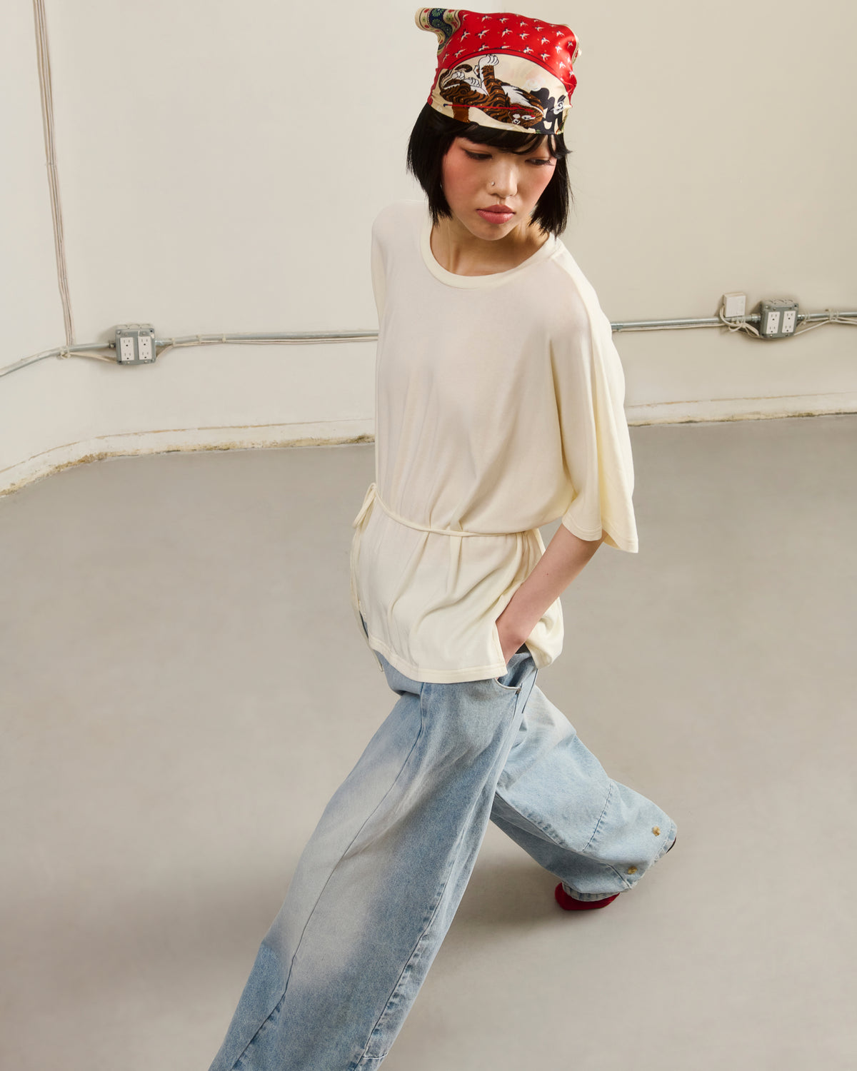 Indoors on a gray floor, a person stands with hands in pockets, wearing a loose cream top, wide-leg light blue jeans, and Sundae School's MINHWA BANDANA silk scarf inspired by Korean folk art.
