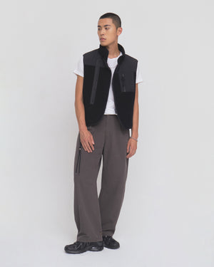A person stands against a plain white background, wearing a white T-shirt, the BLACK MOMO REVERSIBLE FLEECE VEST by Sundae School, loose brown pants, and black shoes. They have short hair and a neutral expression, with one hand relaxed by their side.