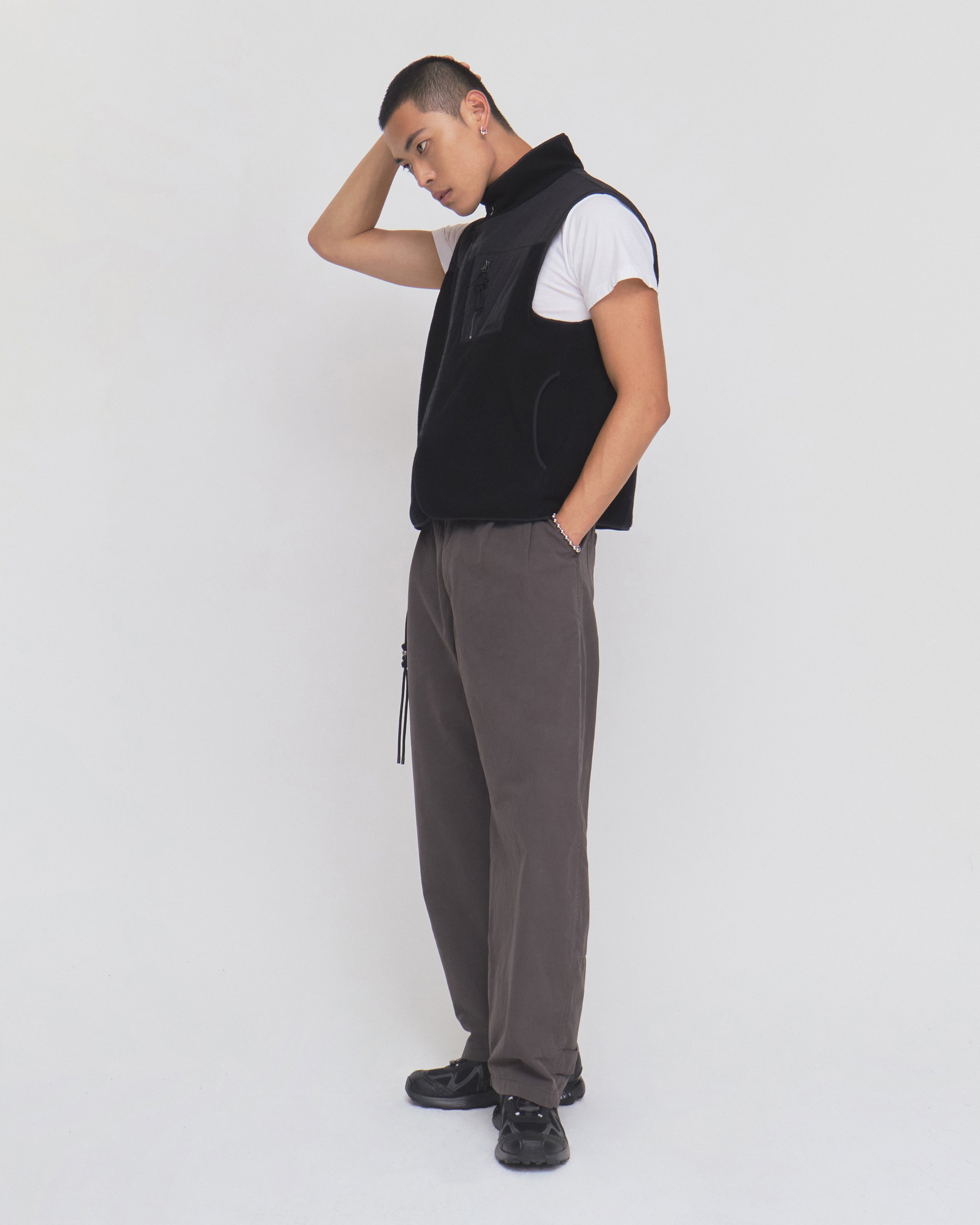 A person stands sideways against a plain white background, wearing a white t-shirt, gray pants, black shoes, and the Sundae School BLACK MOMO REVERSIBLE FLEECE VEST, with one hand behind their head and the other in their pocket.