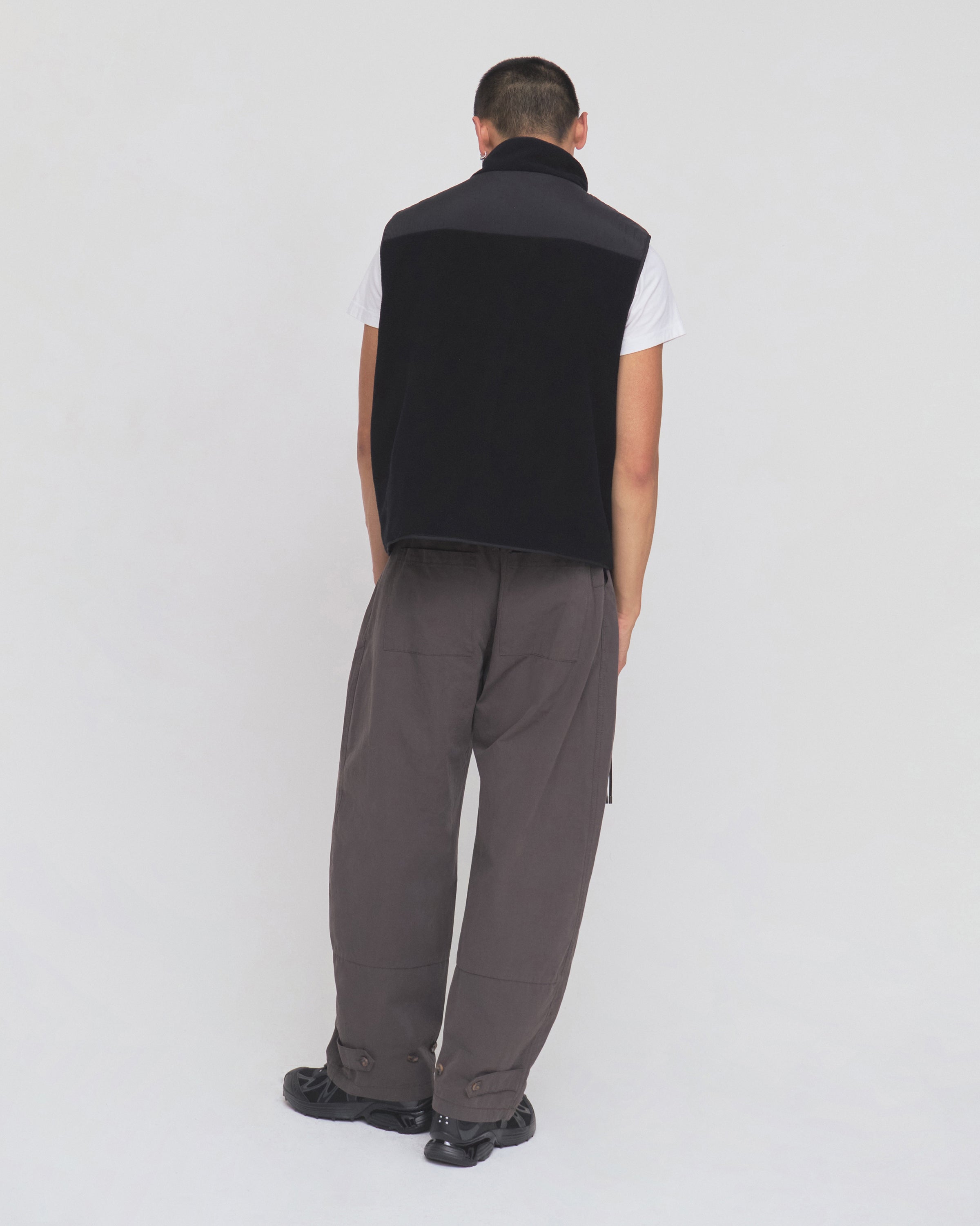 A person stands facing away against a plain white background, wearing the Sundae School BLACK MOMO REVERSIBLE FLEECE VEST over a white T-shirt, paired with loose gray pants and black shoes.