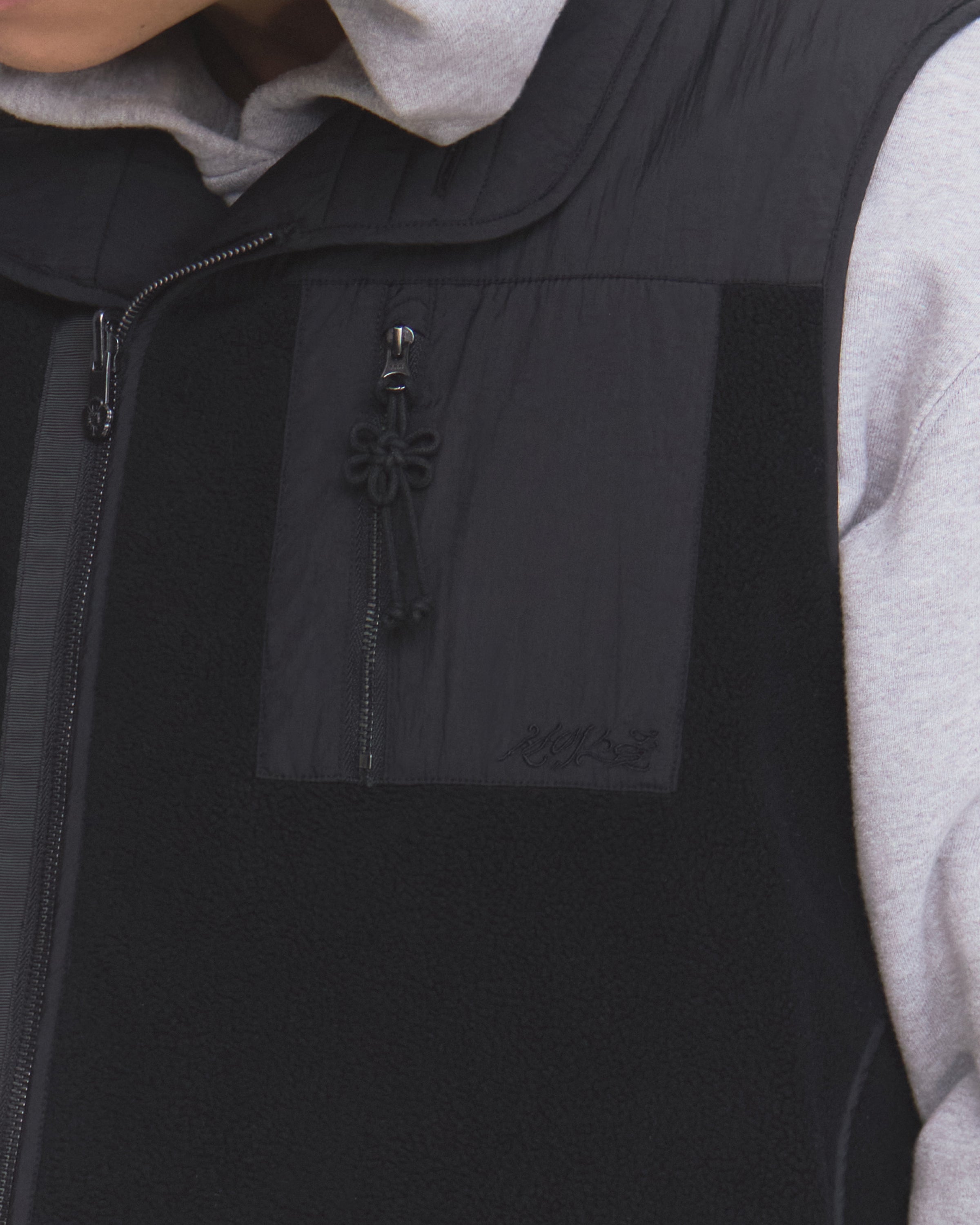 A person wears a light gray hooded sweatshirt under the Sundae School BLACK MOMO REVERSIBLE FLEECE VEST, which features a decorative knotted pull cord on the chest pocket.