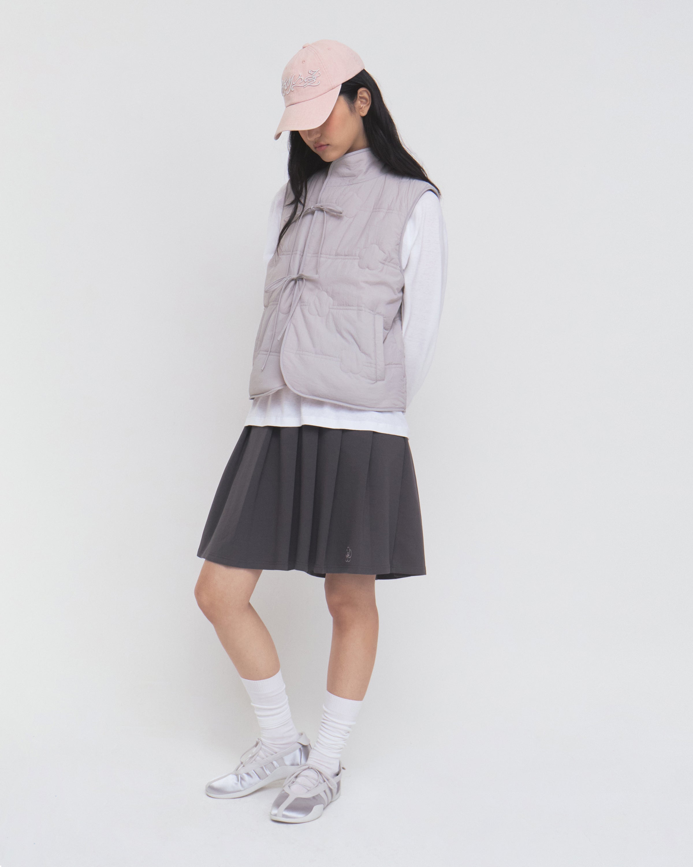 A person wearing the Sundae School BABY PINK MOMO REVERSIBLE FLEECE VEST over a white long-sleeve shirt, paired with a light pink cap, dark pleated skirt, white socks, and sneakers stands against a plain white background, looking downward.