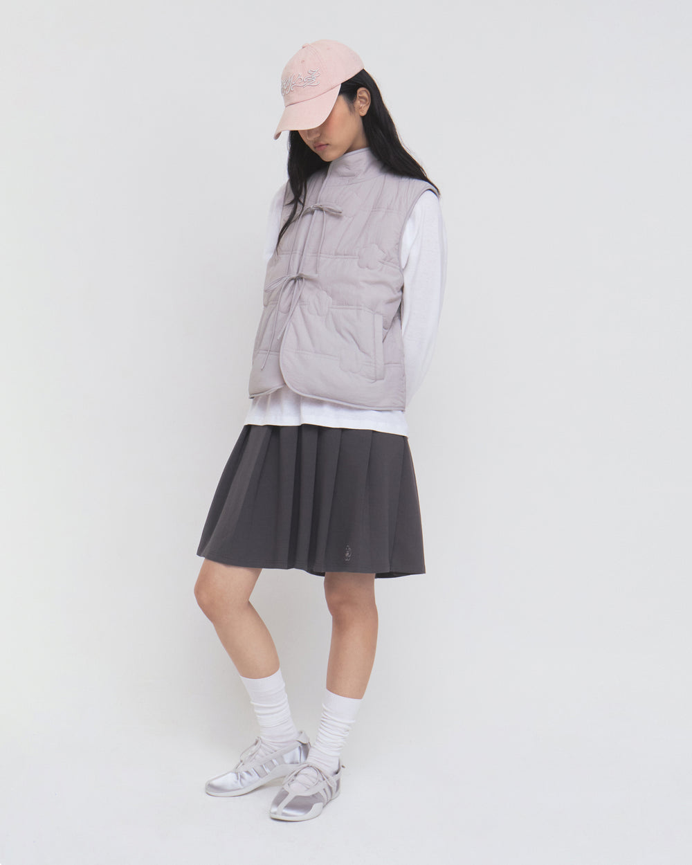 A person wearing the Sundae School BABY PINK MOMO REVERSIBLE FLEECE VEST over a white long-sleeve shirt, paired with a light pink cap, dark pleated skirt, white socks, and sneakers stands against a plain white background, looking downward.
