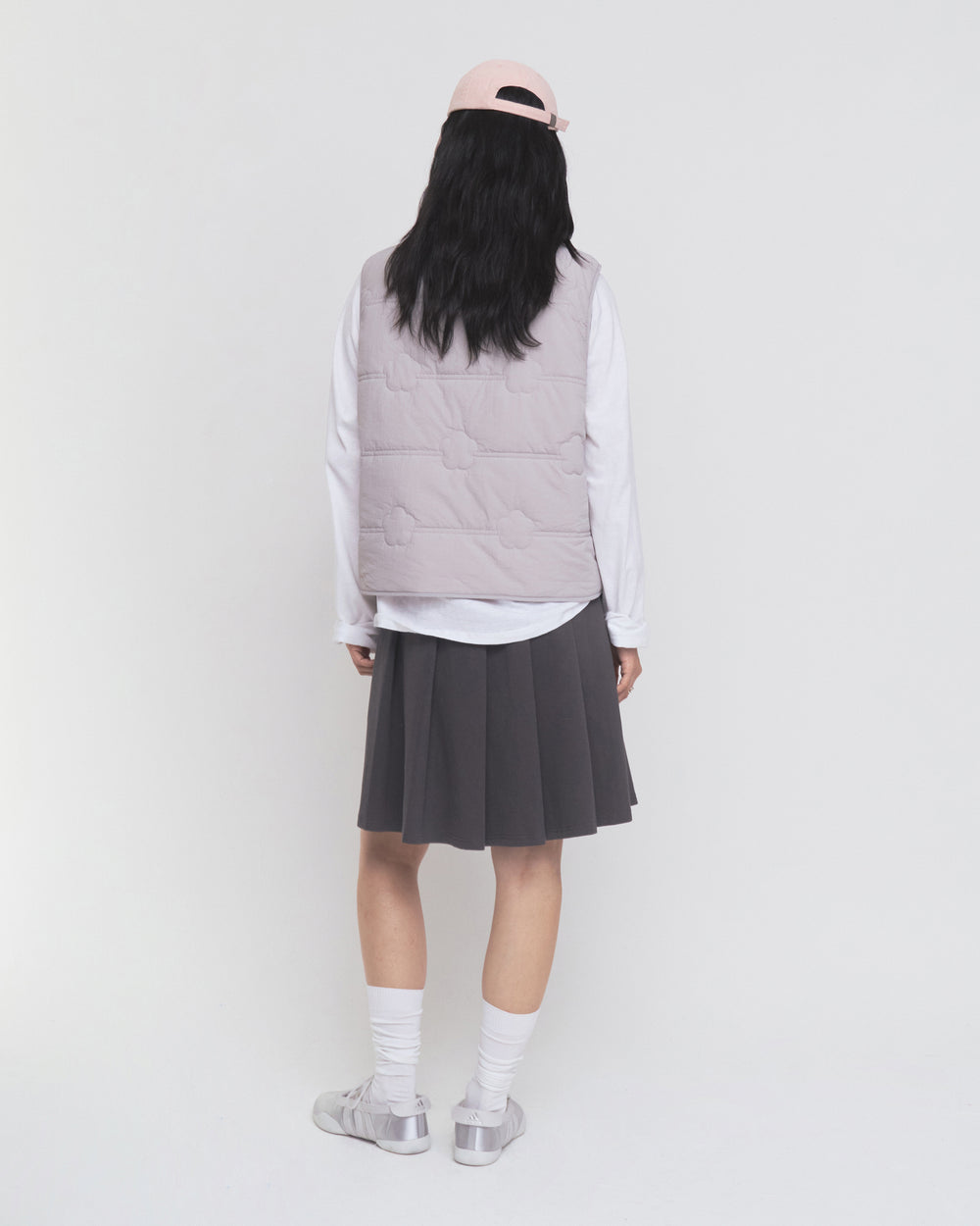 A person stands facing away against a plain white background, wearing a pale pink cap, the Sundae School BABY PINK MOMO REVERSIBLE FLEECE VEST, a white long-sleeve shirt, a dark pleated skirt, white socks, and silver sneakers.