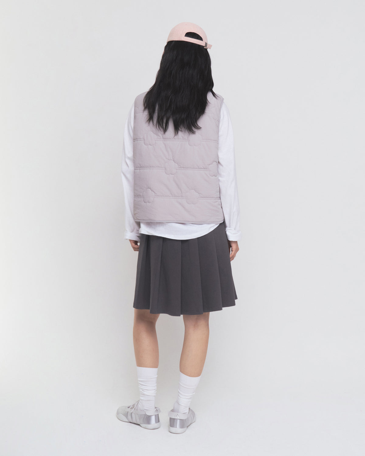A person stands facing away against a plain white background, wearing a pale pink cap, the Sundae School BABY PINK MOMO REVERSIBLE FLEECE VEST, a white long-sleeve shirt, a dark pleated skirt, white socks, and silver sneakers.