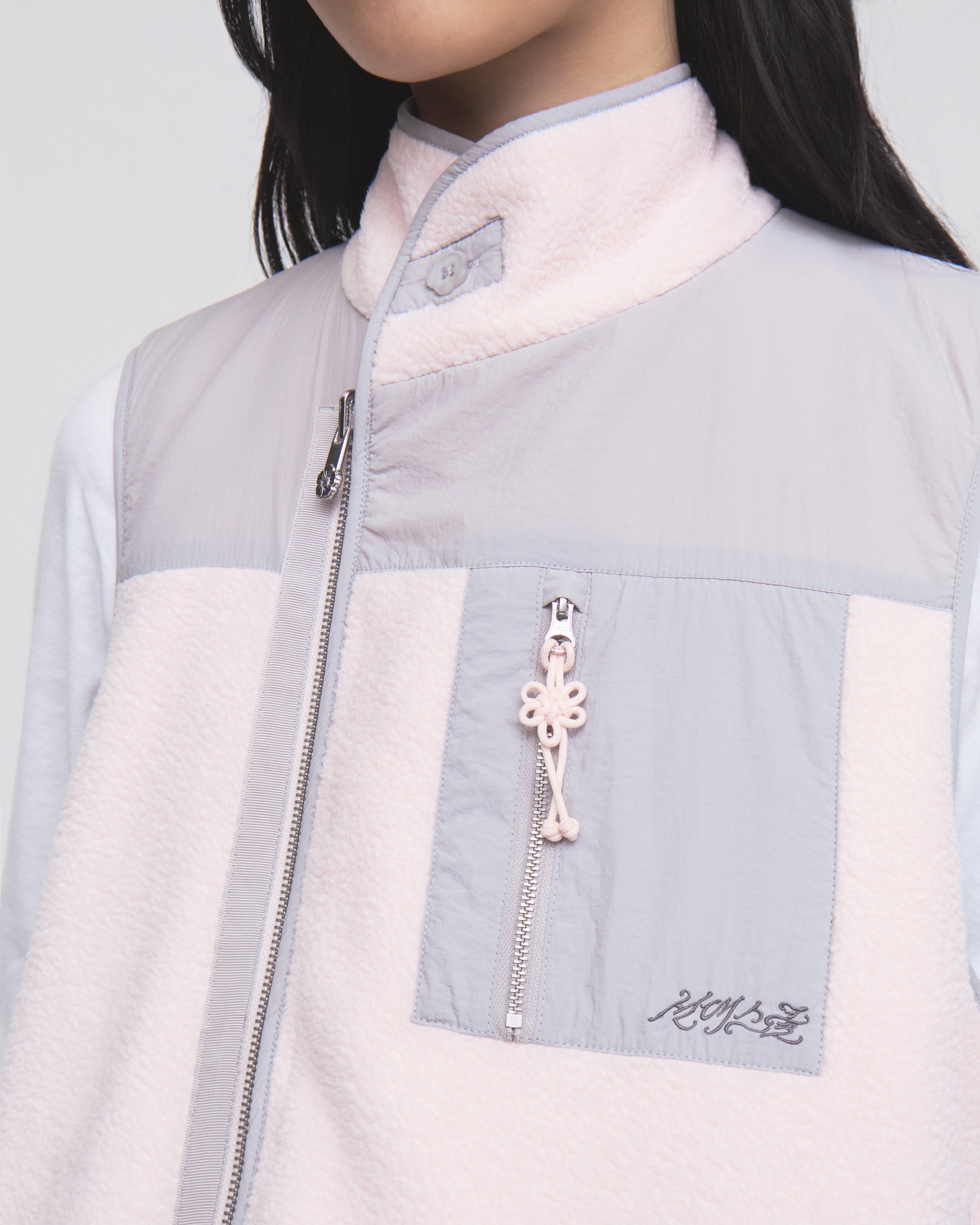 A person is shown from the lower face to the upper torso wearing the Sundae School BABY PINK MOMO REVERSIBLE FLEECE VEST in light pink and gray, which features a zippered chest pocket with an attached pink keychain and embroidered text below the pocket.