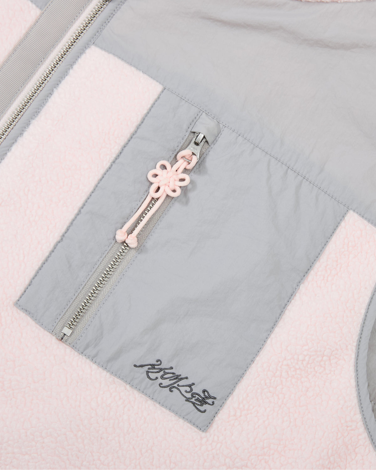 Close-up of the Sundae School BABY PINK MOMO REVERSIBLE FLEECE VEST, featuring light pink fleece, a gray fabric pocket, a zipper with a pink knot pull, and black stylized embroidery near the pocket.