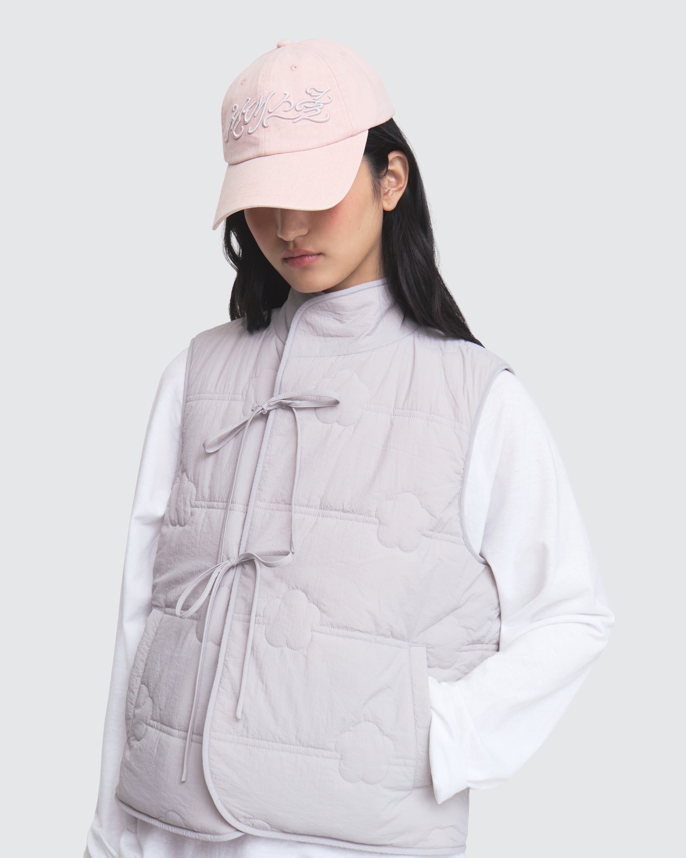 A person wearing the Sundae School BABY PINK MOMO REVERSIBLE FLEECE VEST and a light pink cap over a white long-sleeve shirt stands against a light gray background, looking down.