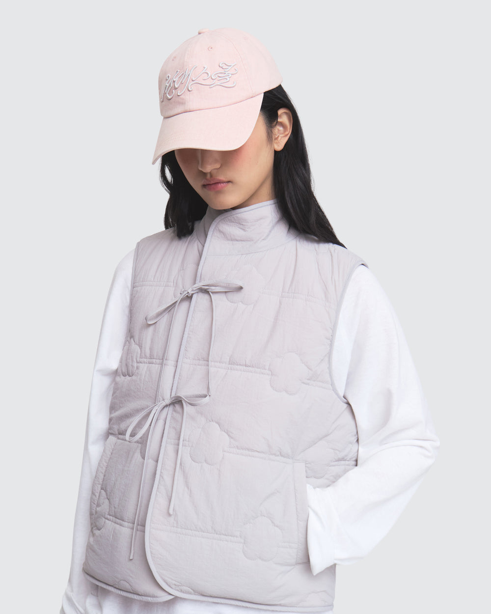 A person wearing the Sundae School BABY PINK MOMO REVERSIBLE FLEECE VEST and a light pink cap over a white long-sleeve shirt stands against a light gray background, looking down.