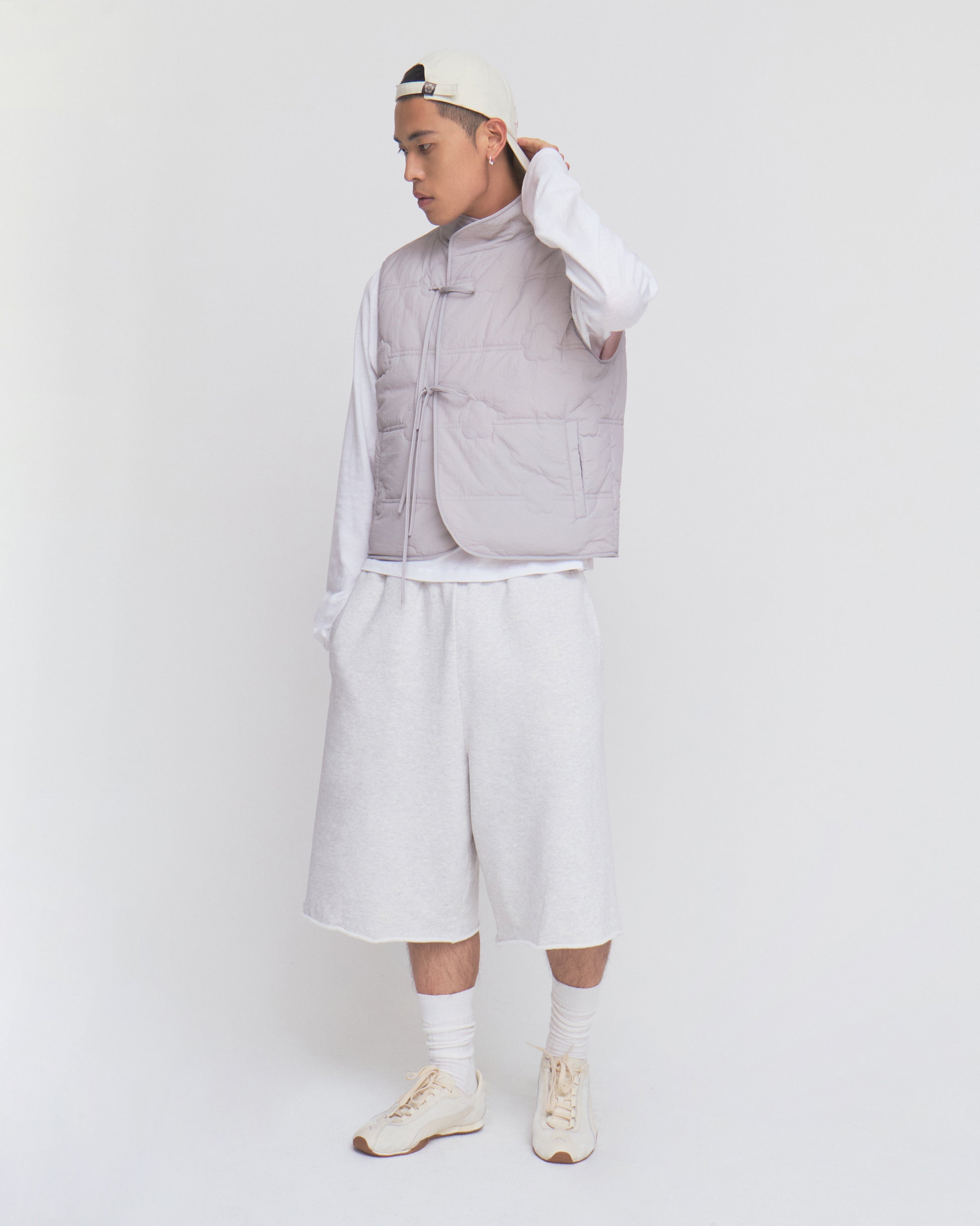 Wearing the BABY PINK MOMO REVERSIBLE FLEECE VEST by Sundae School over a white long-sleeve shirt, loose white shorts, and a backward white cap, this person stands against a plain white background in cream sneakers and white socks, looking to the side.