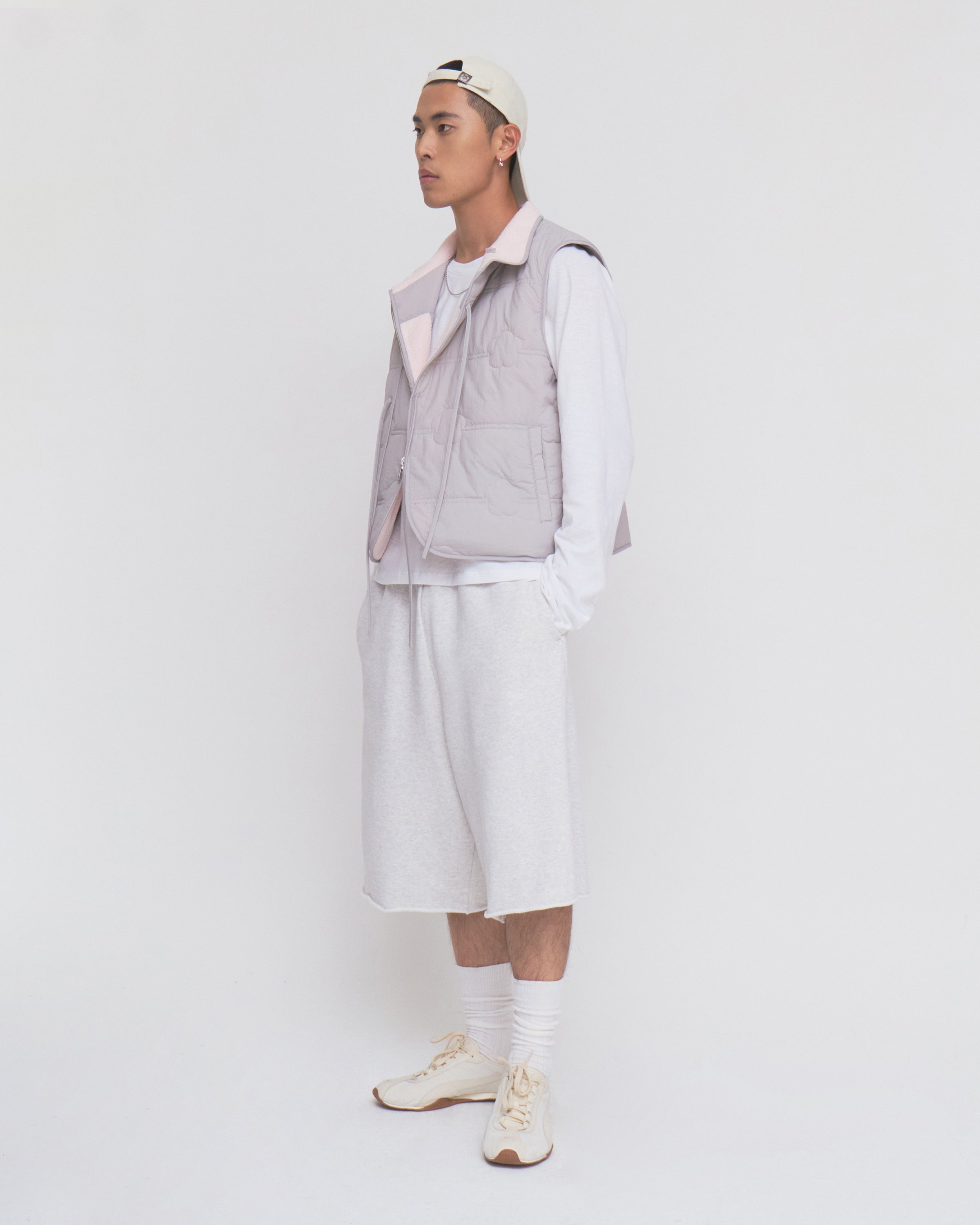 A person in a white cap, BABY PINK MOMO REVERSIBLE FLEECE VEST by Sundae School, white long-sleeve shirt, baggy shorts, socks, and sneakers stands against a plain white background, facing left with a neutral expression.
