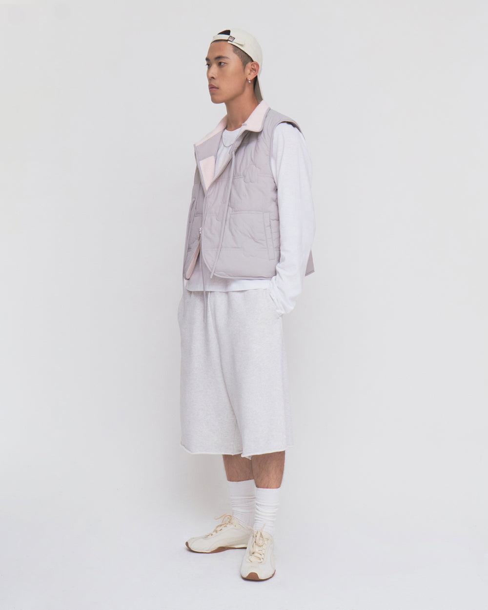 A person in a white cap, BABY PINK MOMO REVERSIBLE FLEECE VEST by Sundae School, white long-sleeve shirt, baggy shorts, socks, and sneakers stands against a plain white background, facing left with a neutral expression.