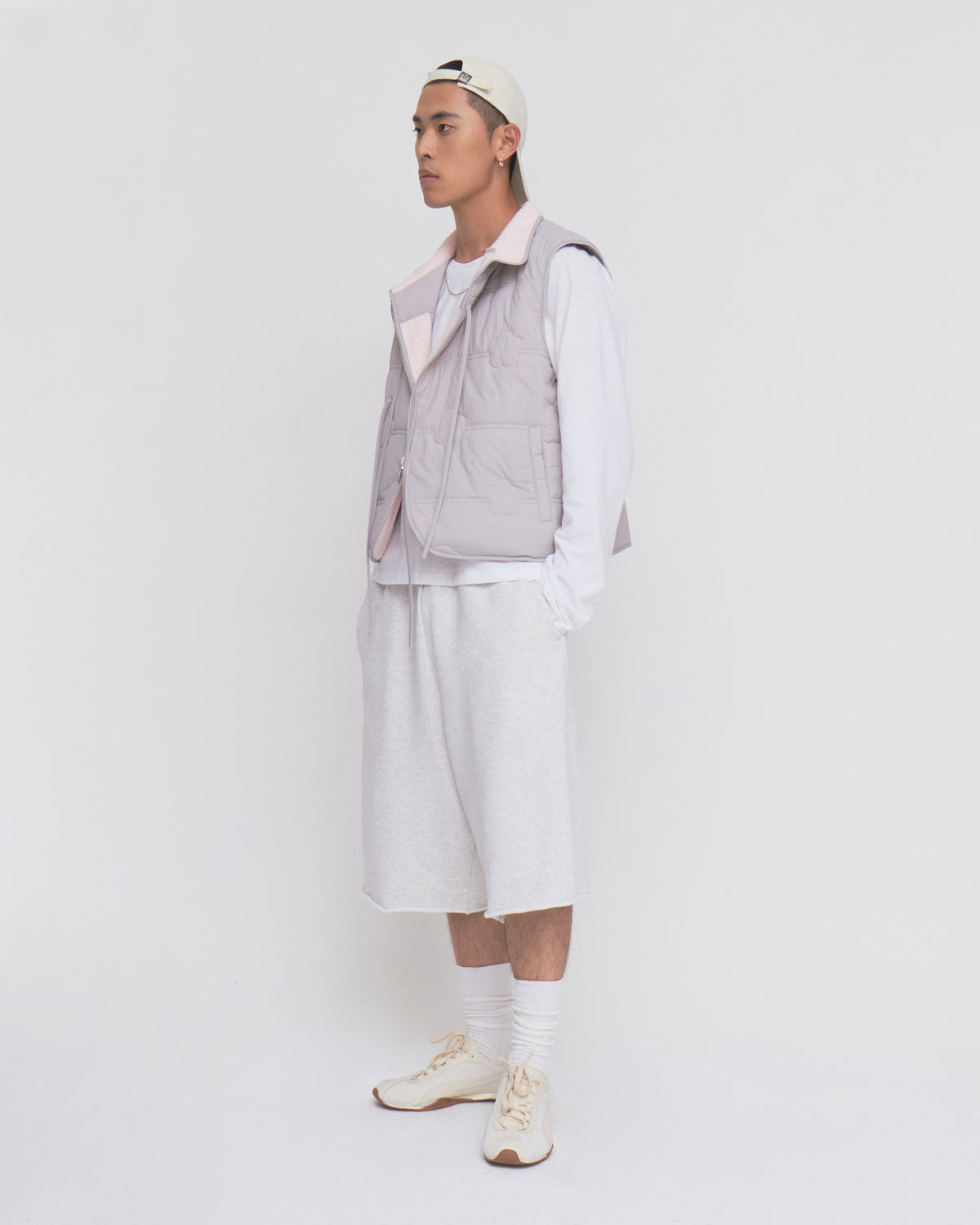 A person in a white cap, BABY PINK MOMO REVERSIBLE FLEECE VEST by Sundae School, white long-sleeve shirt, baggy shorts, socks, and sneakers stands against a plain white background, facing left with a neutral expression.