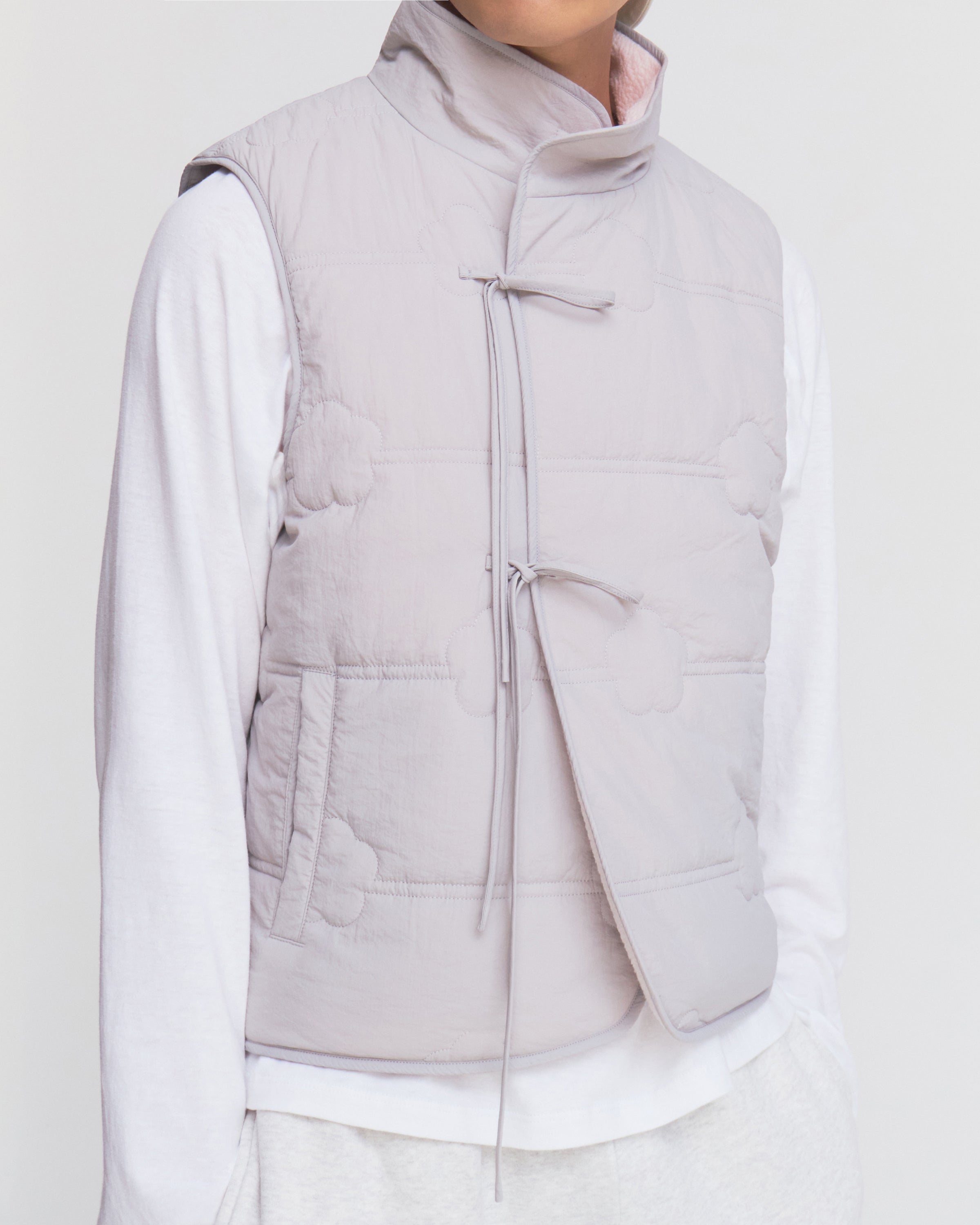 A person wears the Sundae School BABY PINK MOMO REVERSIBLE FLEECE VEST, a light gray quilted piece with subtle cloud-shaped stitching and a side tie closure, layered over a long-sleeve white shirt. The head is partially out of frame against a plain background.