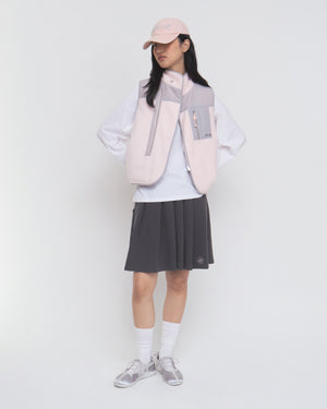 A person stands against a plain white background wearing the Sundae School BABY PINK MOMO REVERSIBLE FLEECE VEST over a white long-sleeve shirt, paired with a light pink cap, dark pleated skirt, white socks, and gray sneakers.