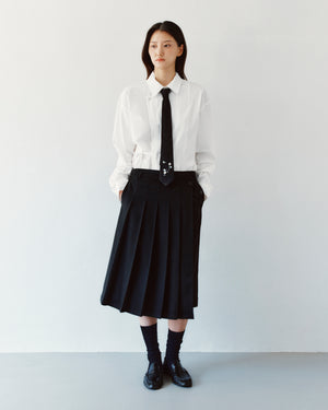 A person wears a white button-down shirt, a thin black necktie with floral embroidery, and a BLACK MOON WRAP MIDI SKIRT featuring an asymmetric pleated design.