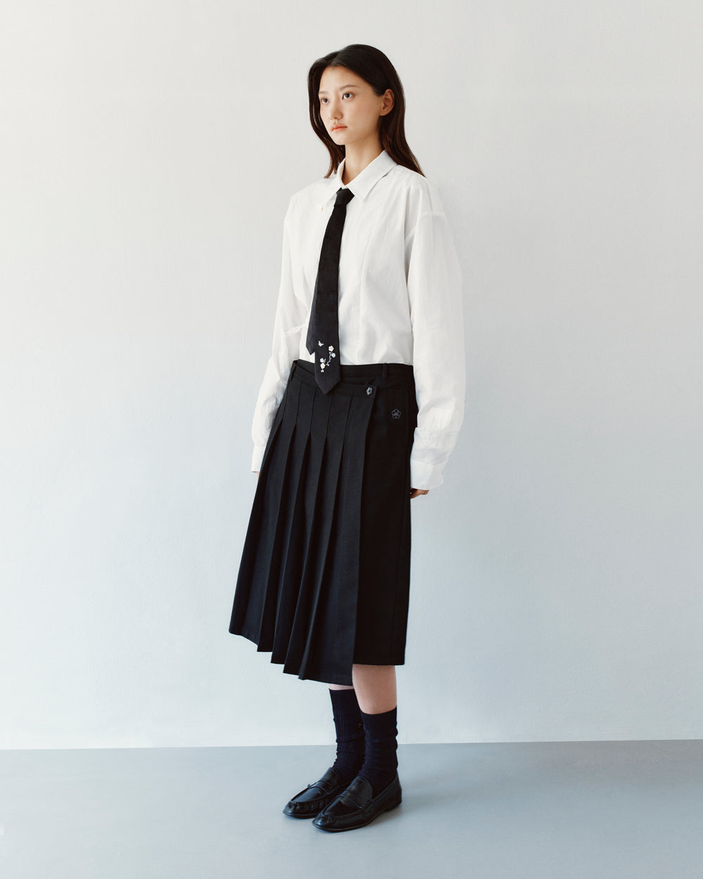 A person wears a white button-down shirt and a thin black necktie with a BLACK MOON WRAP MIDI SKIRT, an asymmetric pleated style featuring an adjustable waist belt.