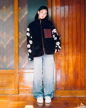 A person stands indoors in front of wooden and frosted glass doors, wearing the Sundae School MUGUNGHWA FLEECE—a black sherpa jacket with floral sleeves inspired by the Korean national flower—paired with loose blue jeans and light-colored shoes. The distinctive MUGUNGHWA motif on the sleeves adds a subtle, elegant touch to the look.