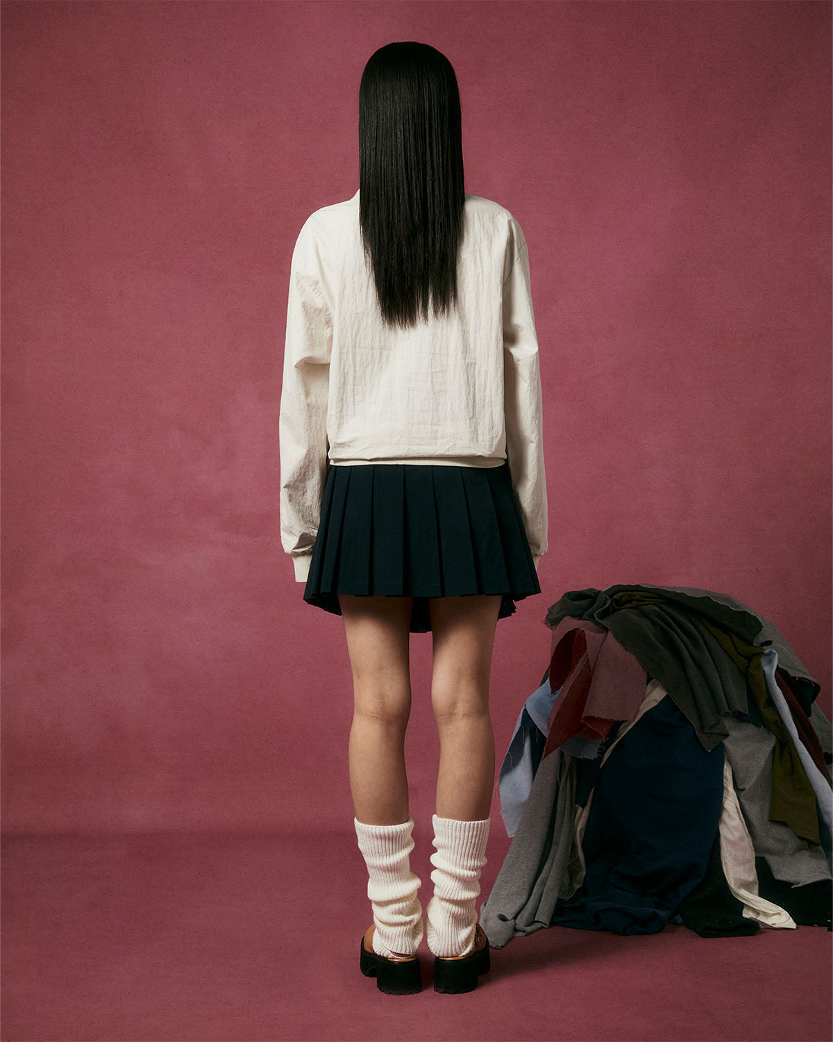 A person with straight black hair faces away against a pink background, wearing Sundae School’s IVORY SHAWL COLLAR ANORAK, a black pleated skirt, white leg warmers, and black sandals. A pile of clothes sits on the floor to the right.