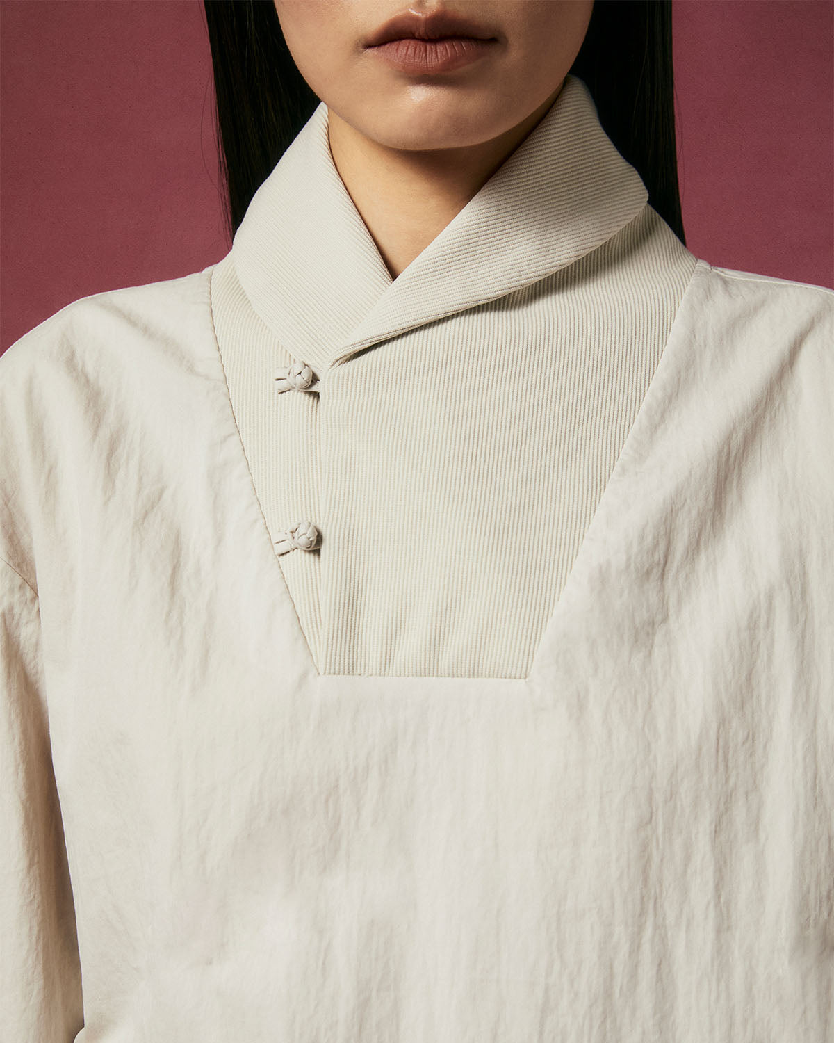 A person wears the Sundae School IVORY SHAWL COLLAR ANORAK, featuring a textured white fabric, high shawl collar, and two knotted buttons, set against a dark pink background with only the lower half of their face visible.