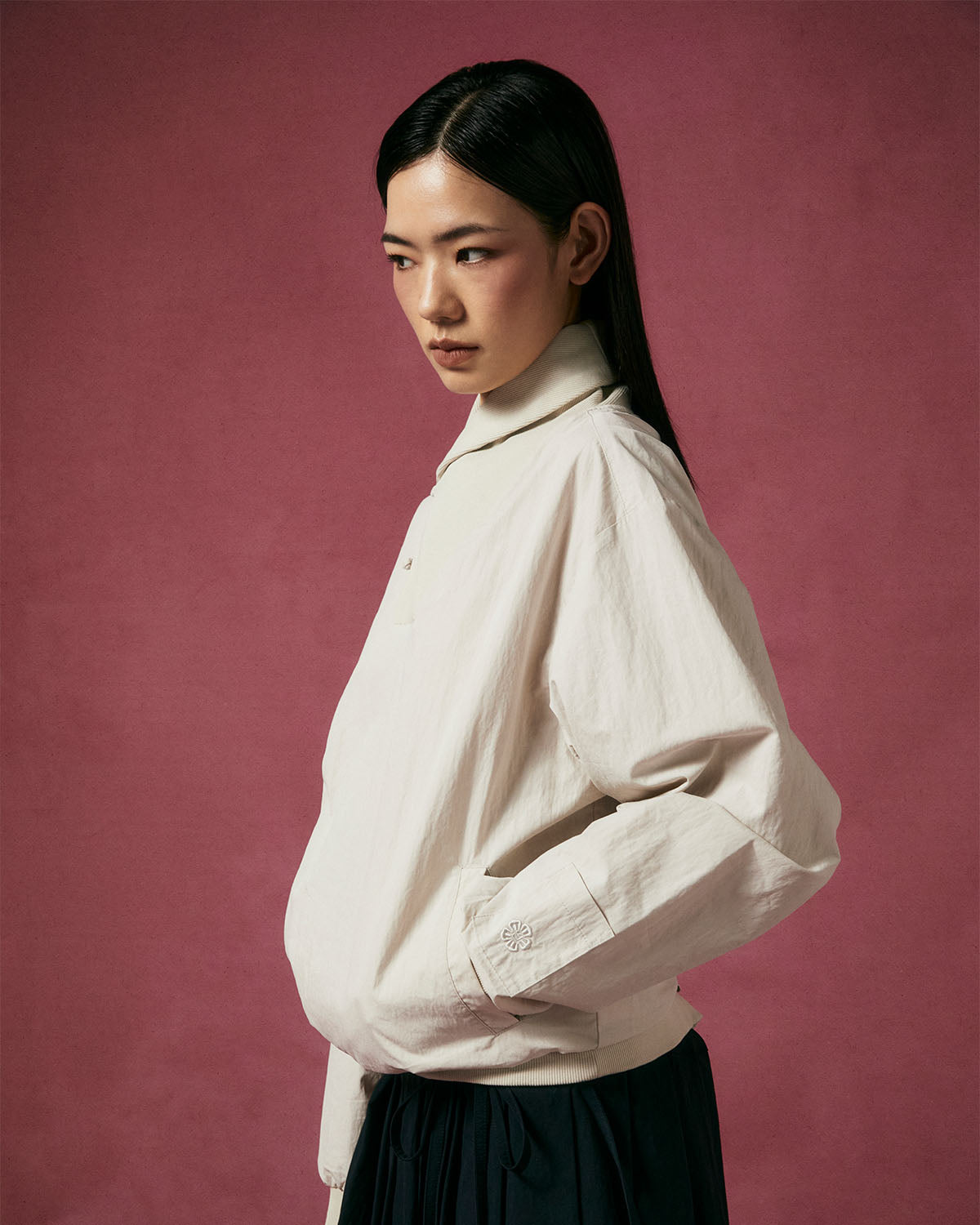 A person with straight black hair, wearing the IVORY SHAWL COLLAR ANORAK by Sundae School and a dark skirt, stands in profile against a plain maroon background.