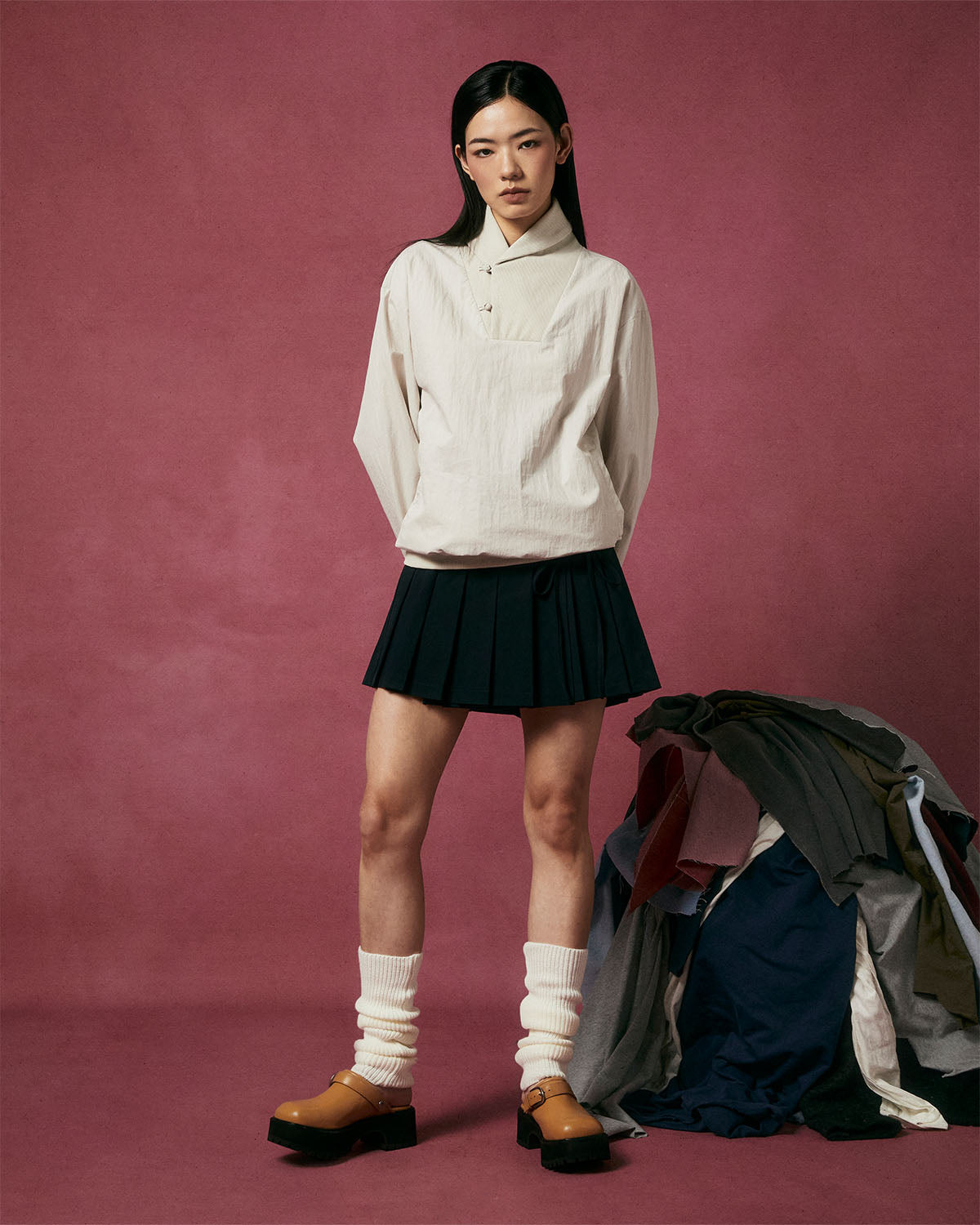 A woman stands against a reddish backdrop, wearing the Sundae School IVORY SHAWL COLLAR ANORAK, a black pleated skirt, white leg warmers, and tan platform shoes. A pile of clothes sits on the floor beside her.