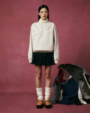 A woman with straight dark hair stands against a pink background, wearing Sundae School’s IVORY SHAWL COLLAR ANORAK, a black pleated skirt, and white leg warmers. A pile of assorted clothing rests on the floor beside her.