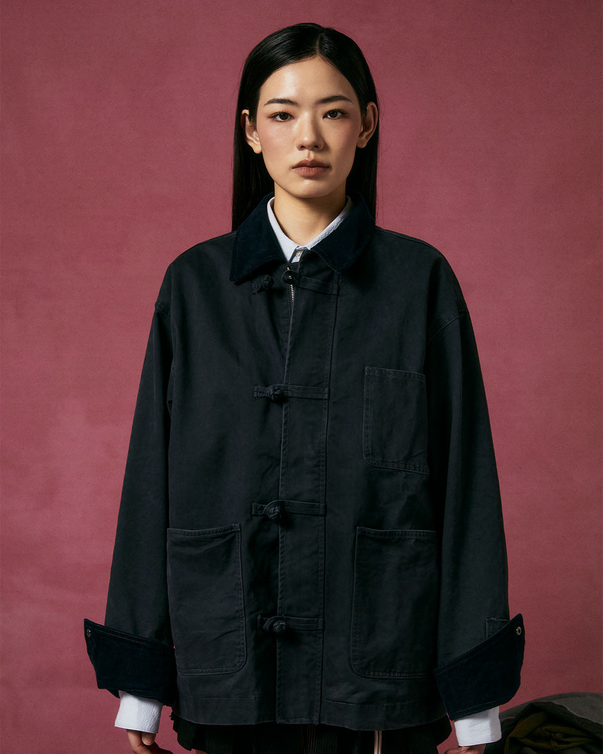 A person with straight black hair wears the Sundae School CHARCOAL KNOT BARN JACKET over a white shirt, standing against a maroon background and looking at the camera with a neutral expression.