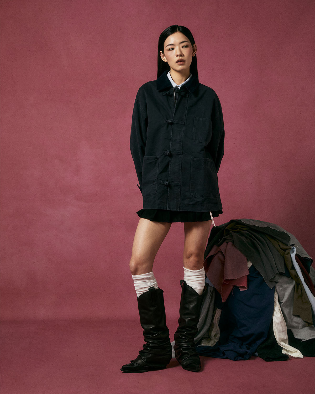 A woman stands before a pink backdrop in Sundae School's CHARCOAL KNOT BARN JACKET, inspired by Joseon military workwear, styled with a short skirt, white socks, black boots, and her hands behind her back near a pile of clothes.