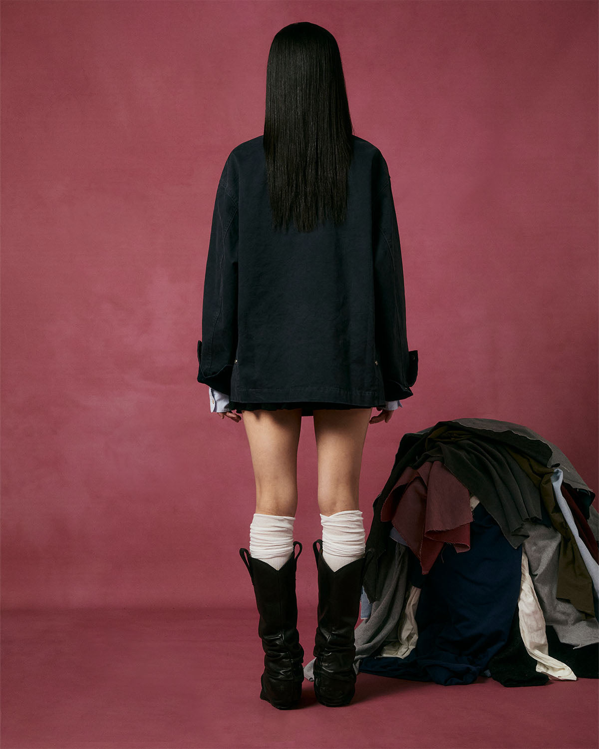A person with long straight hair faces away, wearing the Sundae School CHARCOAL KNOT BARN JACKET styled with workwear flair, plus white legwarmers and black boots. A pile of clothes sits to their right against a pink backdrop.