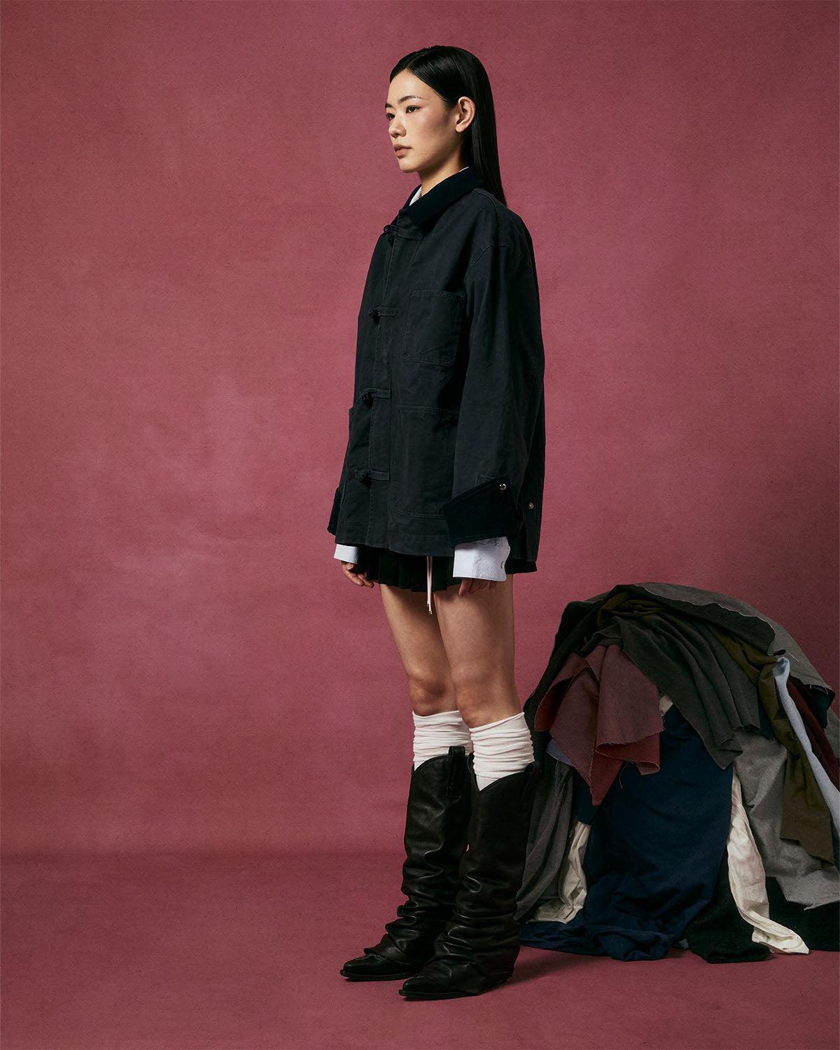A woman in profile stands against a pink background wearing the Sundae School CHARCOAL KNOT BARN JACKET, styled with a white shirt, black shorts, white leg warmers, and black boots. Assorted clothing is piled on the floor beside her.