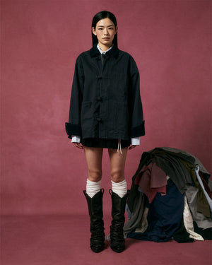 A woman stands against a dark pink backdrop, wearing the CHARCOAL KNOT BARN JACKET by Sundae School over a white shirt and short black skirt, paired with white knee-high socks and black slouchy boots. A pile of assorted clothes lies on the floor beside her.