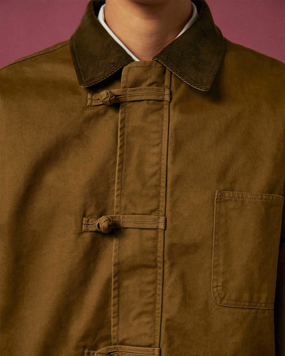 A close-up of a person wearing the TAN KNOT BARN JACKET by Sundae School, featuring a corduroy collar, toggle buttons, and chest pocket. The mauve background complements the workwear style; the person's face is partially out of frame.