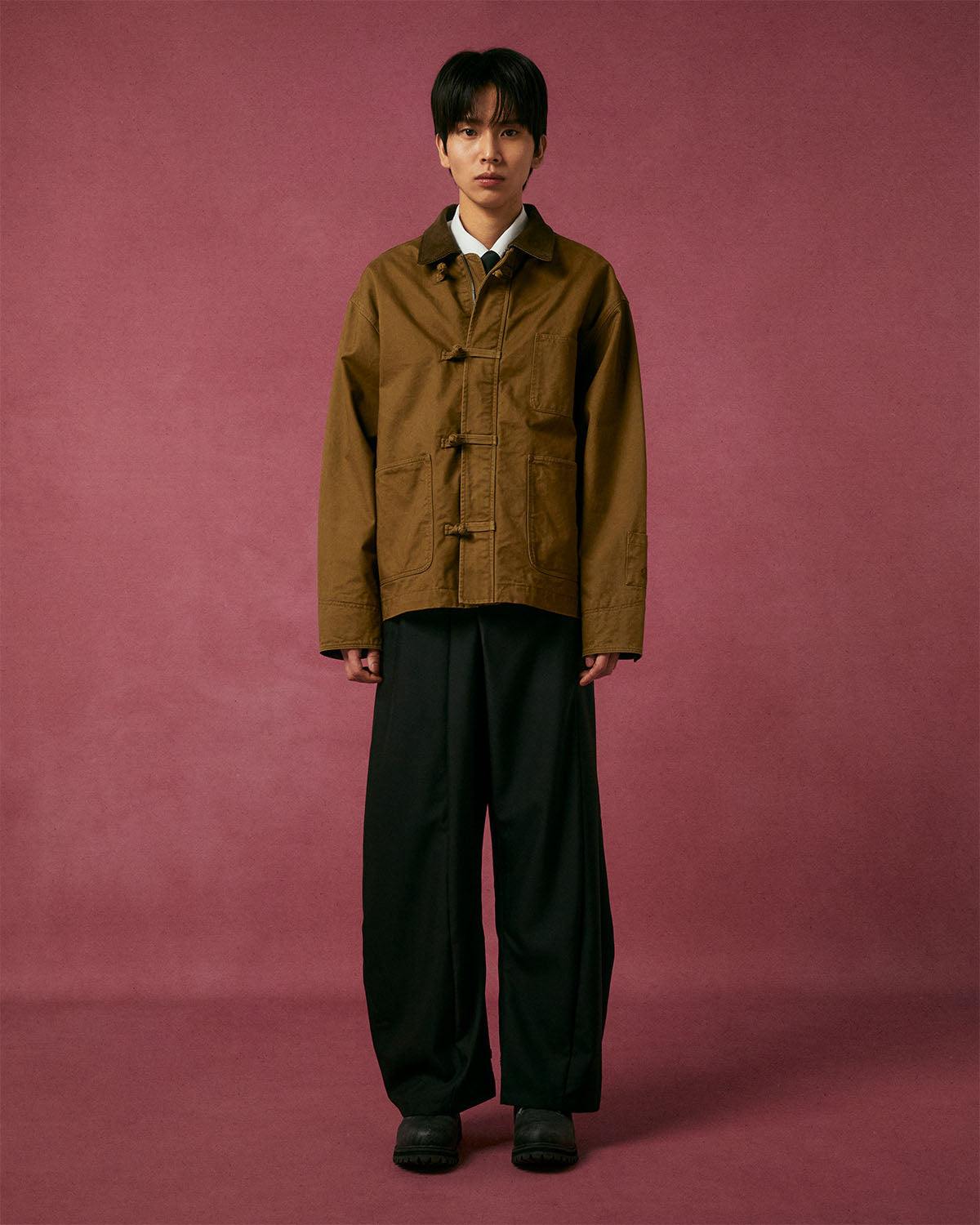 A person stands against a plain maroon backdrop, wearing Sundae School’s TAN KNOT BARN JACKET over a white collared shirt and loose black trousers, paired with black shoes.