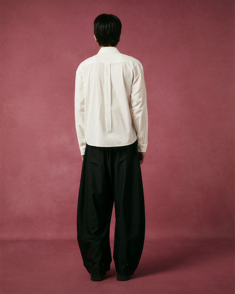 A person stands facing away from the camera against a maroon background, wearing Sundae School's IVORY TIE GORUM SHIRT and loose-fitting black pants.