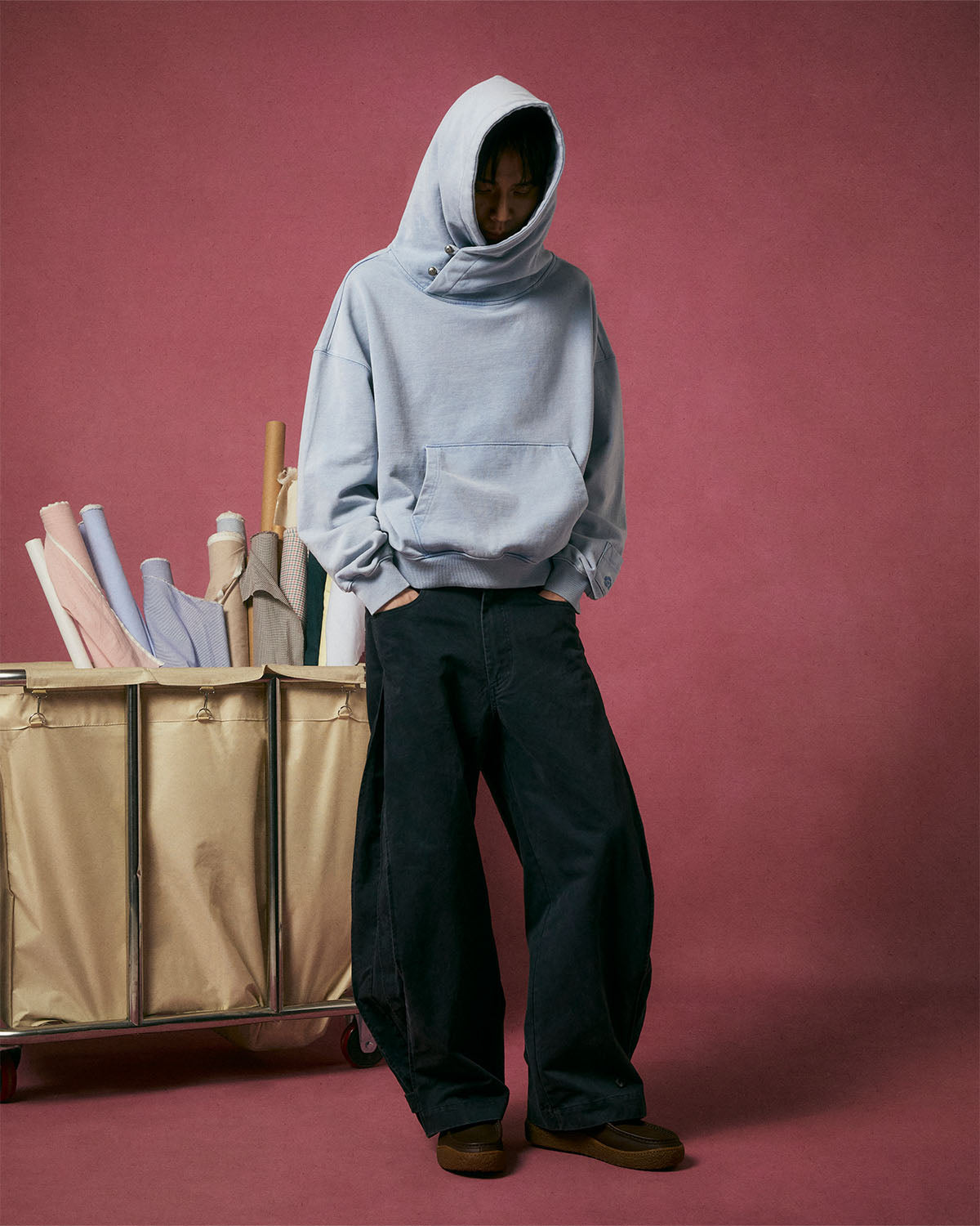 Wearing an oversized light gray hoodie with the hood up and Sundae School BLACK ACCORDION CHINO PANTS, a person stands before a pink backdrop next to a cart filled with rolls of fabric.