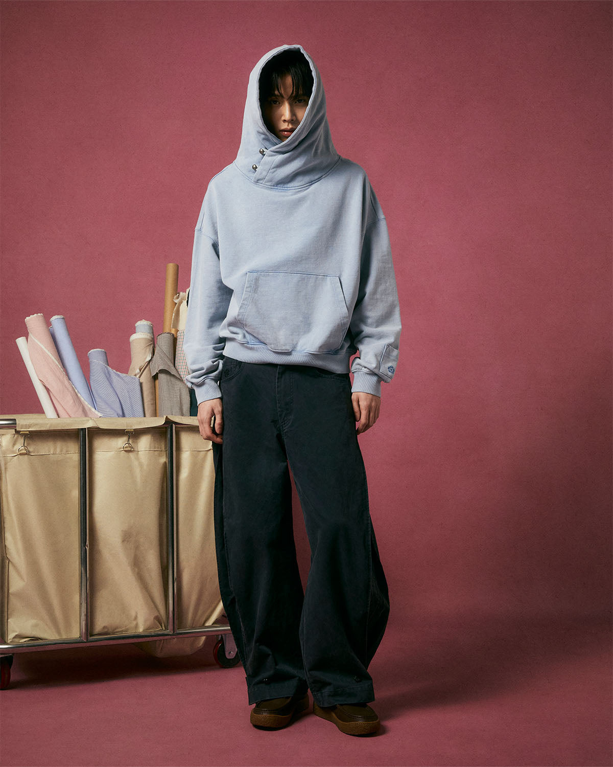 A person in a light gray hooded sweatshirt and Sundae School BLACK ACCORDION CHINO PANTS stands by a beige cart with fabric rolls, against a pink backdrop, wearing brown shoes.