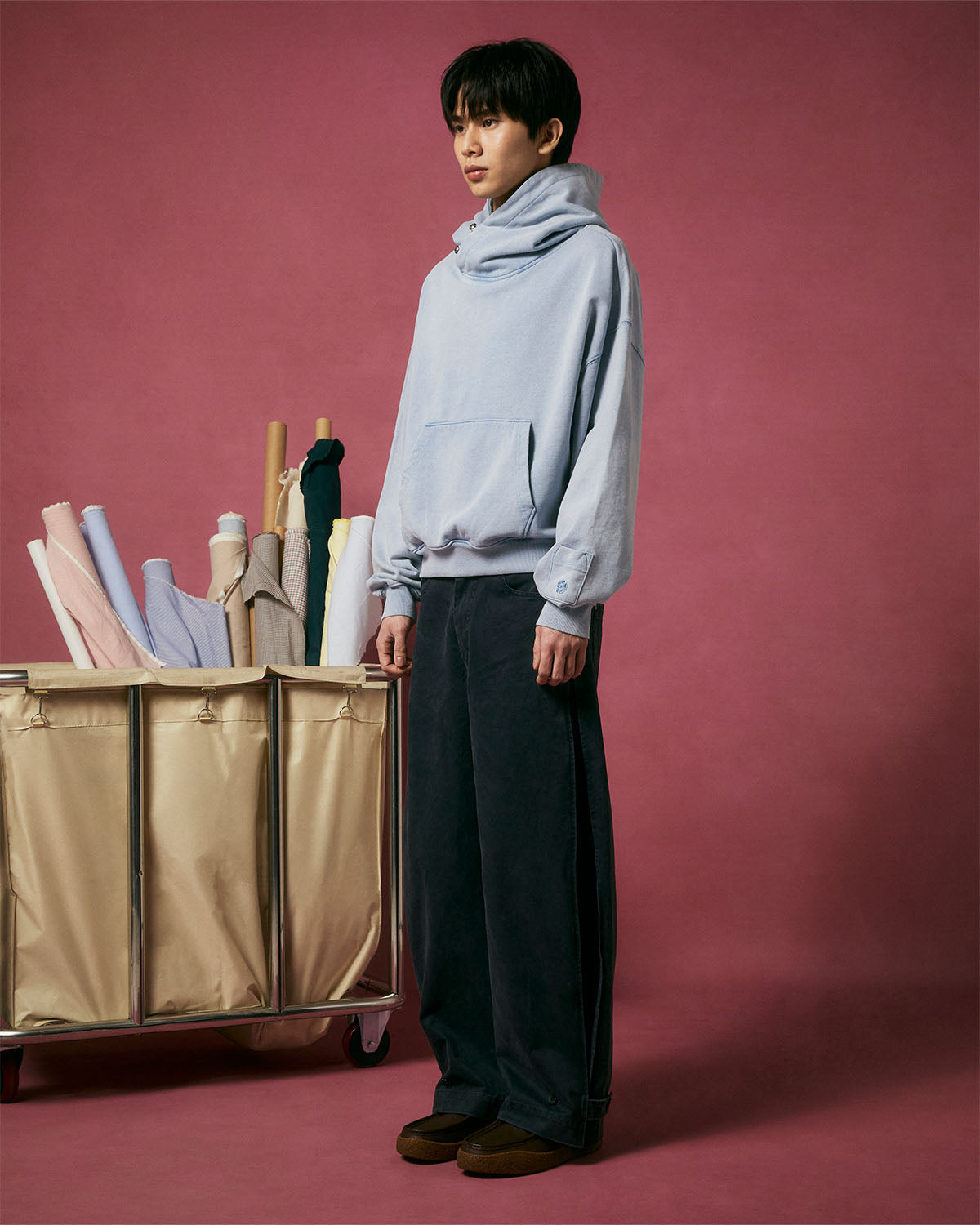 A person in a light blue hoodie and Sundae School BLACK ACCORDION CHINO PANTS stands beside a beige cart stacked with rolled-up fabric bolts, set against a pink background.