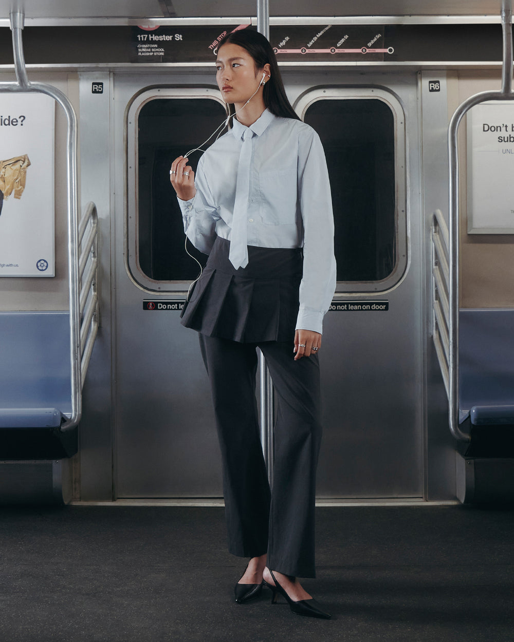 A woman stands in a subway car wearing Sundae School’s NECKTIE SHIRT, styled with a removable tie, paired with a black pleated skirt over black pants and black heels. Listening to earphones, she gazes thoughtfully to the side.