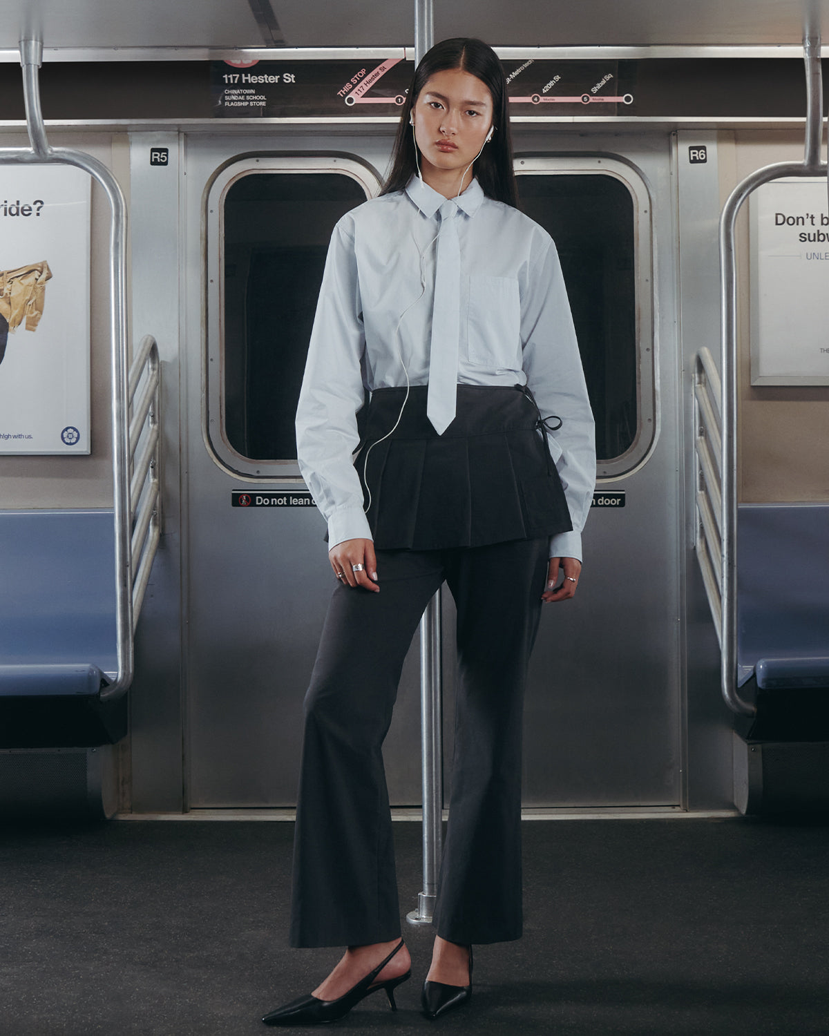 A woman stands on a subway train wearing the Sundae School NECKTIE SHIRT—a relaxed light blue shirt with a removable tie—paired with a black skirt, dark pants, and black heels. She faces forward; subway seats and signage are visible behind her.
